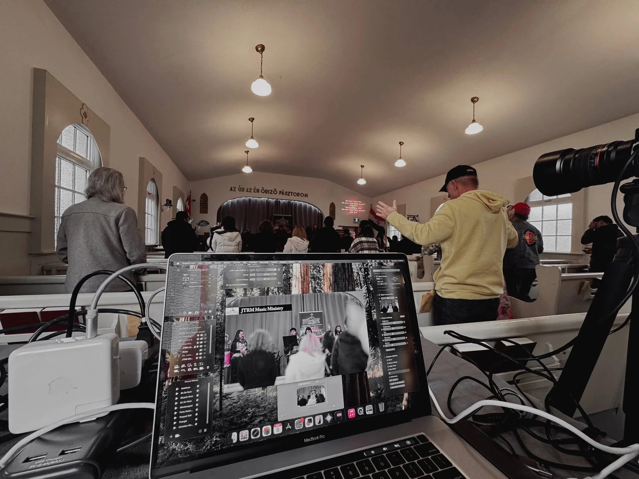 People attending a church service or concert in a church hall with a laptop and camera set up in the foreground.