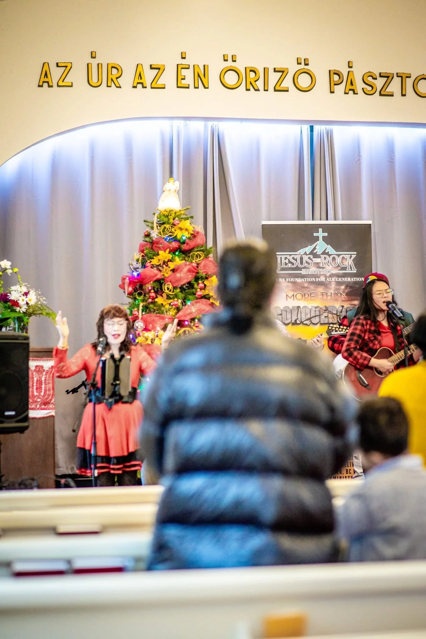 People performing on stage with Christmas decorations, including a tree and flowers, during a church event.