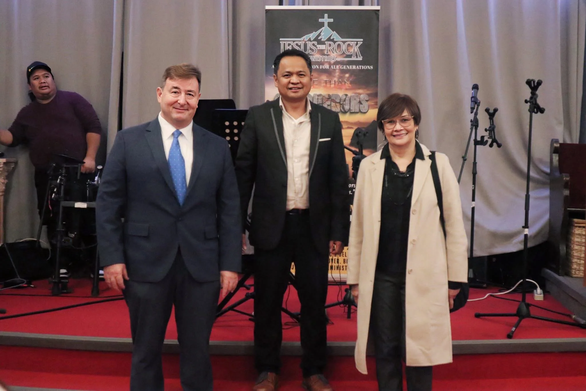 Three people standing in front of a stage with musical instruments and a banner that reads 'Jesus is the Rock Ministries.' The group includes two men and one woman, dressed in business attire, smiling at the camera. A fourth person is seen in the bac