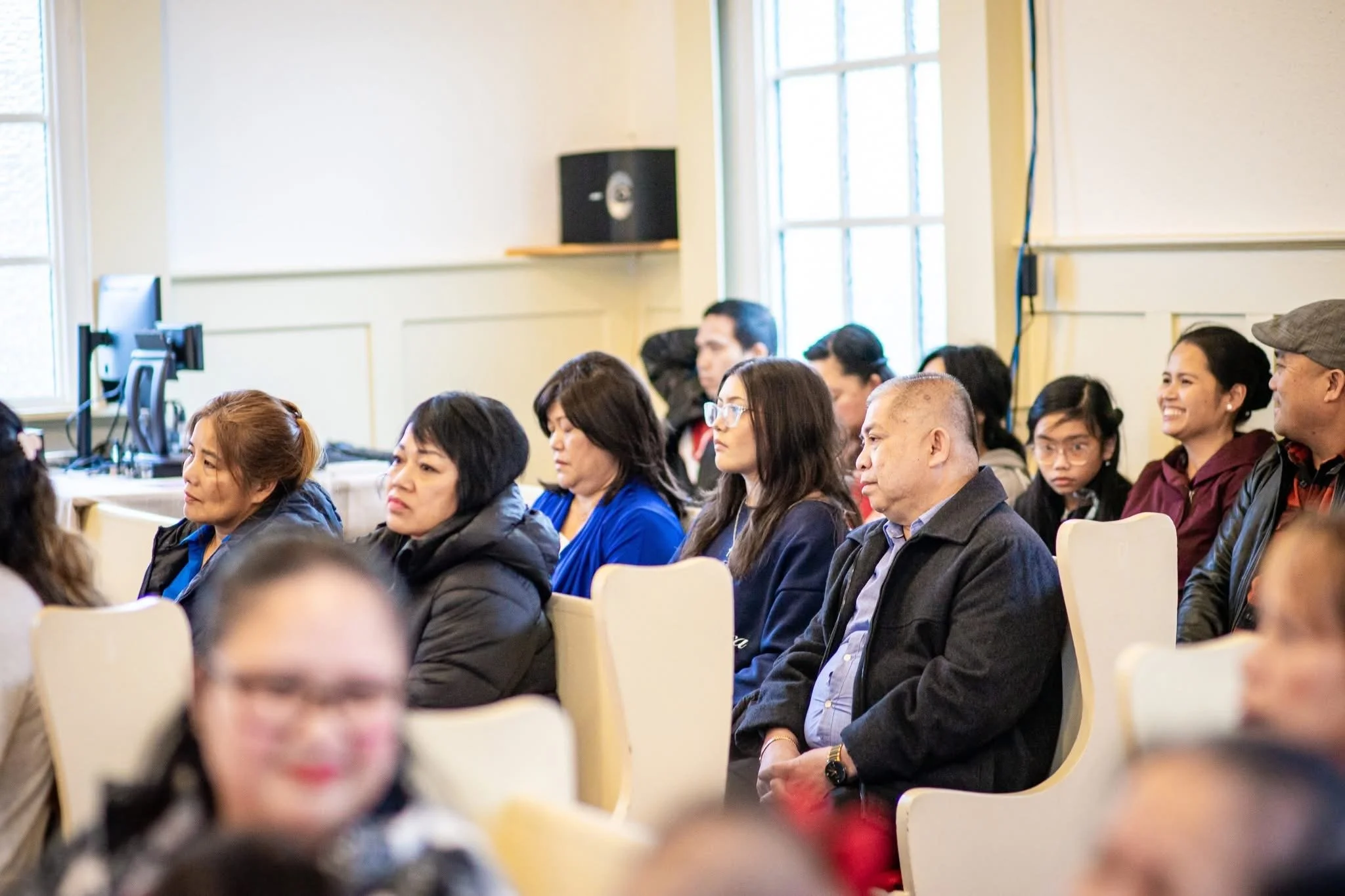 A group of diverse people seated in an indoor conference or seminar room, attentively listening, with some wearing glasses and jackets, and a computer and speaker in the background.