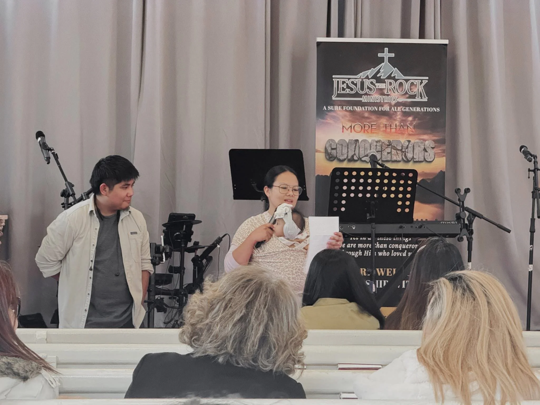 A woman with glasses and a microphone standing on stage, reading from paper, with a man standing nearby, in front of a musical keyboard, microphones, and a banner that says 'Jesus the Rock Ministries' and 'More Than Conquerors' behind them, in an ind