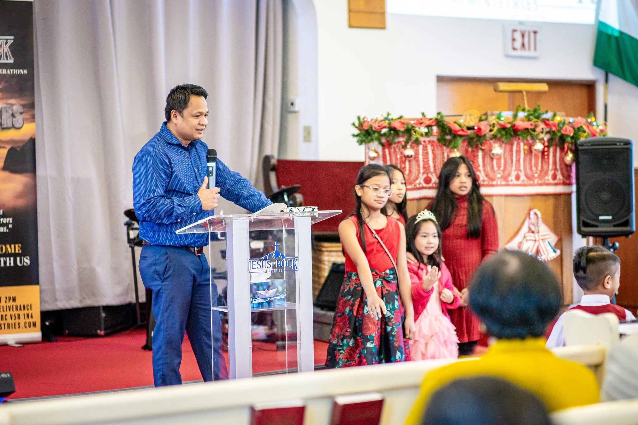 Man speaking at a church podium with children's choir standing beside him, decorated with Christmas-themed decor.