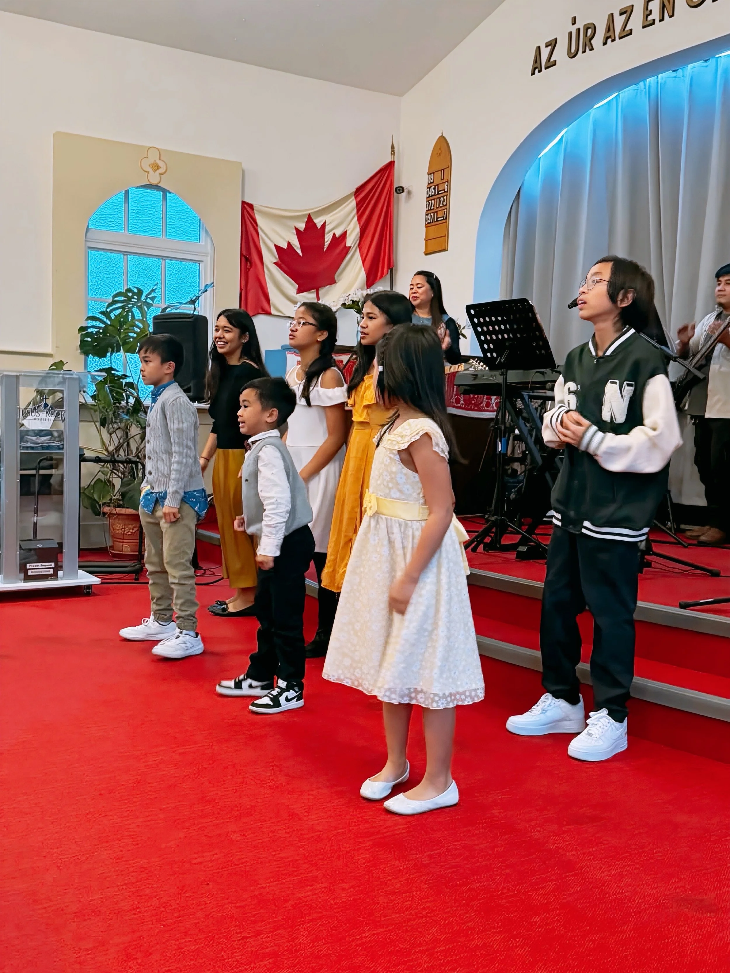 Children and adults standing on stage in a church, with a Canadian flag and a window in the background, during a musical performance or ceremony.