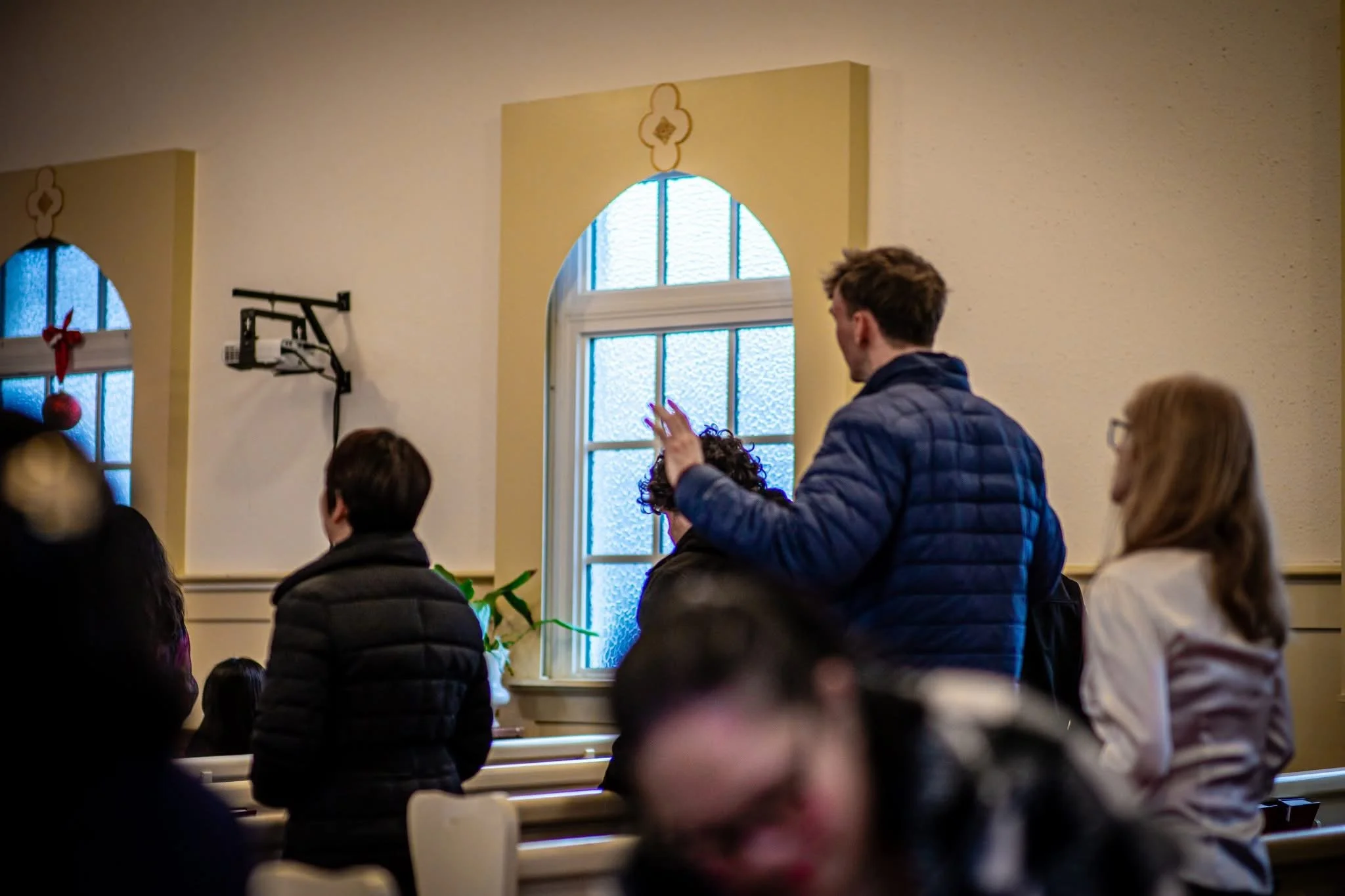A man standing in a church or chapel, raising his hand, engaged in conversation or prayer, with other seated and standing people around him, in a room with stained glass windows and decorative wall accents.