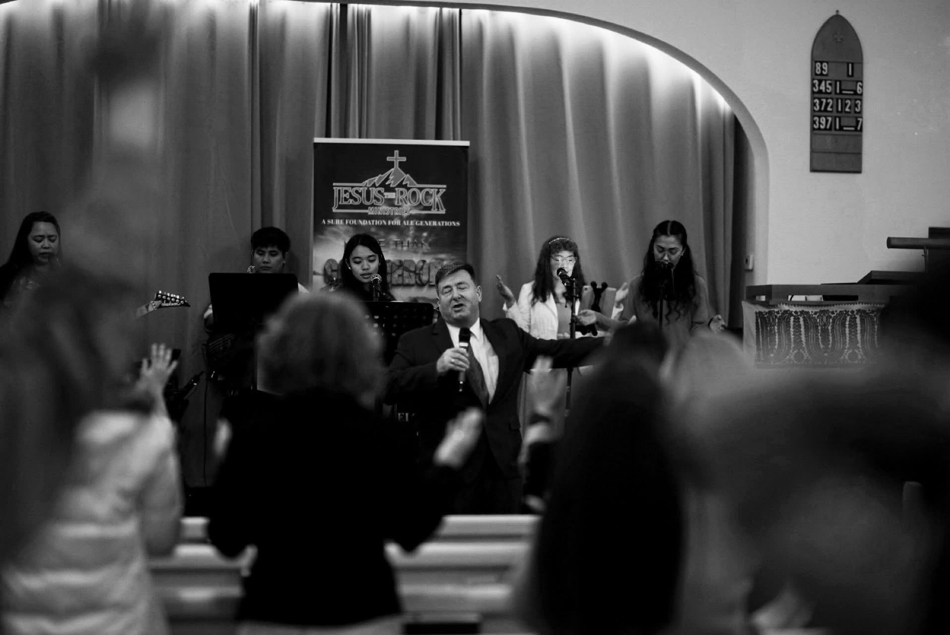 A man in a suit leading a worship service with a microphone in his hand, standing in front of a group of women singing and playing music in a church. The church has curtains and a hymn board visible, and a banner that reads "Jesus Rock Ministry."
