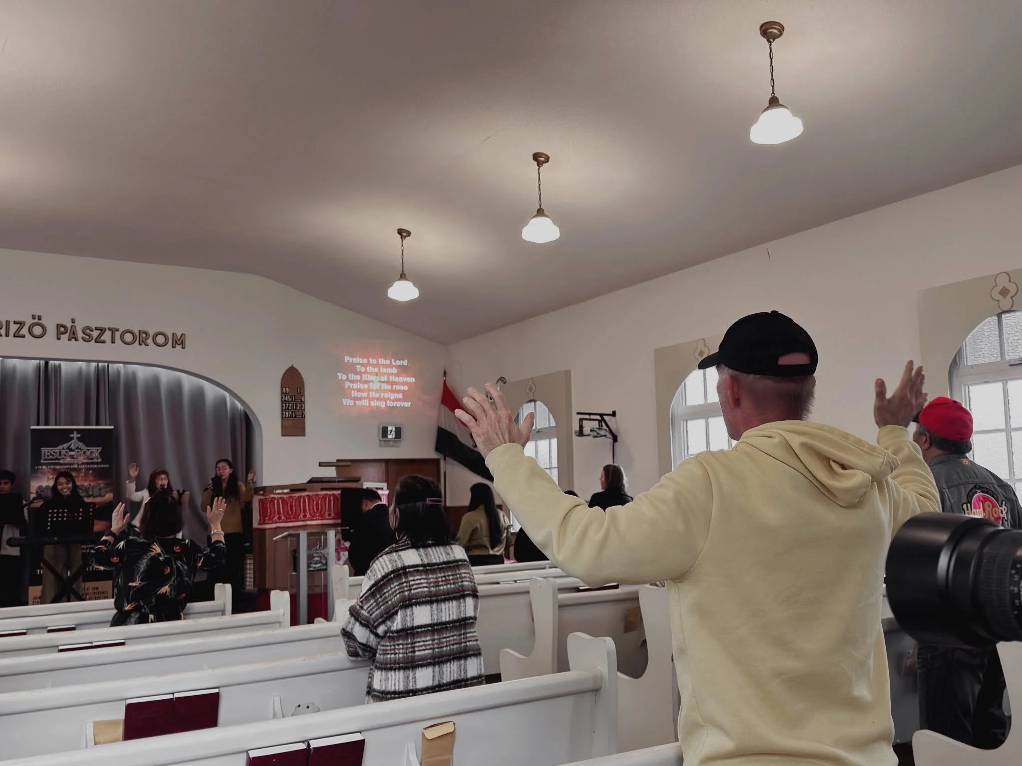 A group of people inside a church or chapel, participating in a religious service. Some are raising their hands during singing or prayer, while others are seated, facing the front where a choir or speakers are performing. The interior has white walls