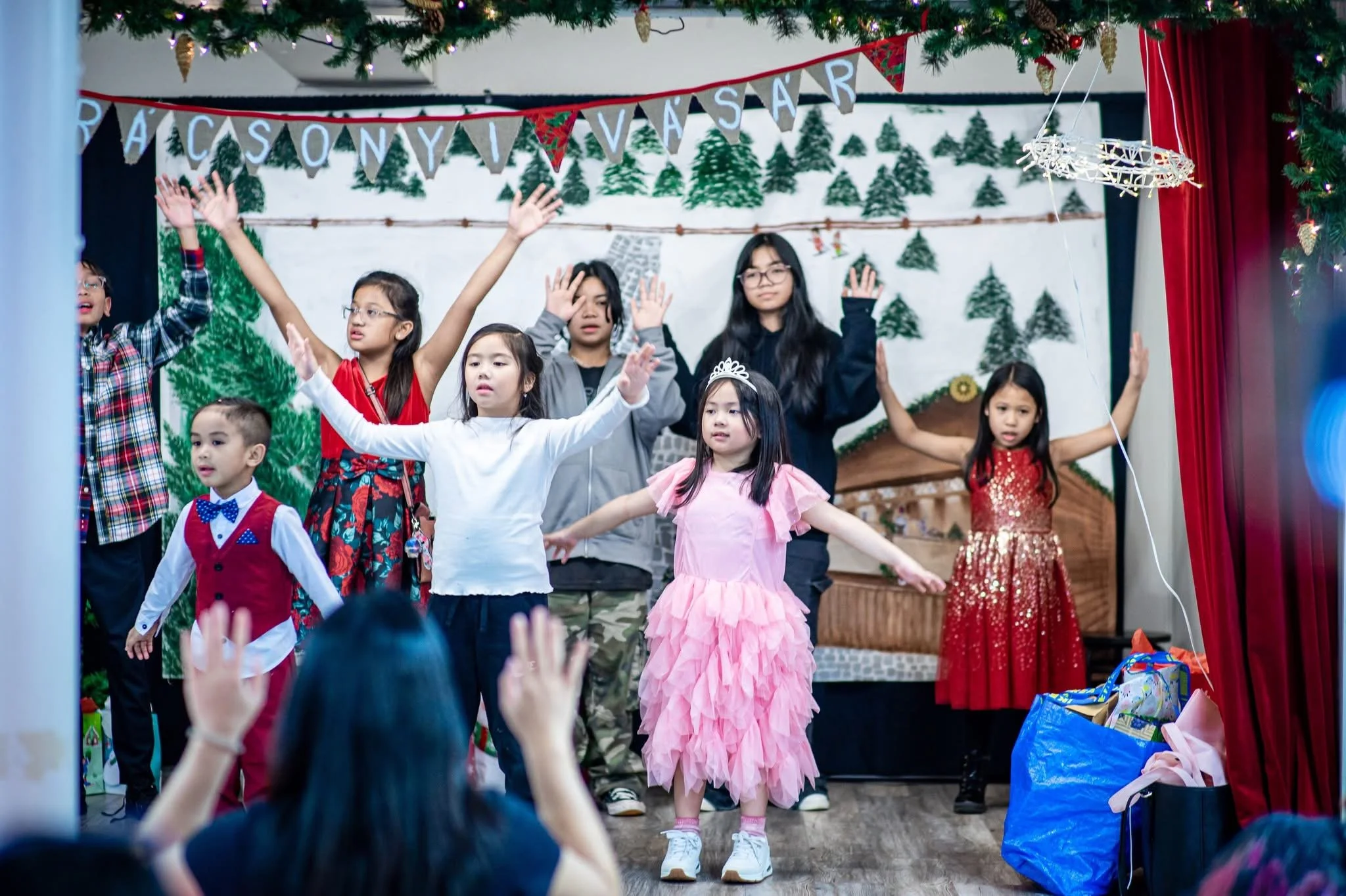 Children performing on stage during a Christmas celebration with festive decorations, including a banner that reads 'PACS A NYI VASA' and a backdrop featuring Christmas trees.
