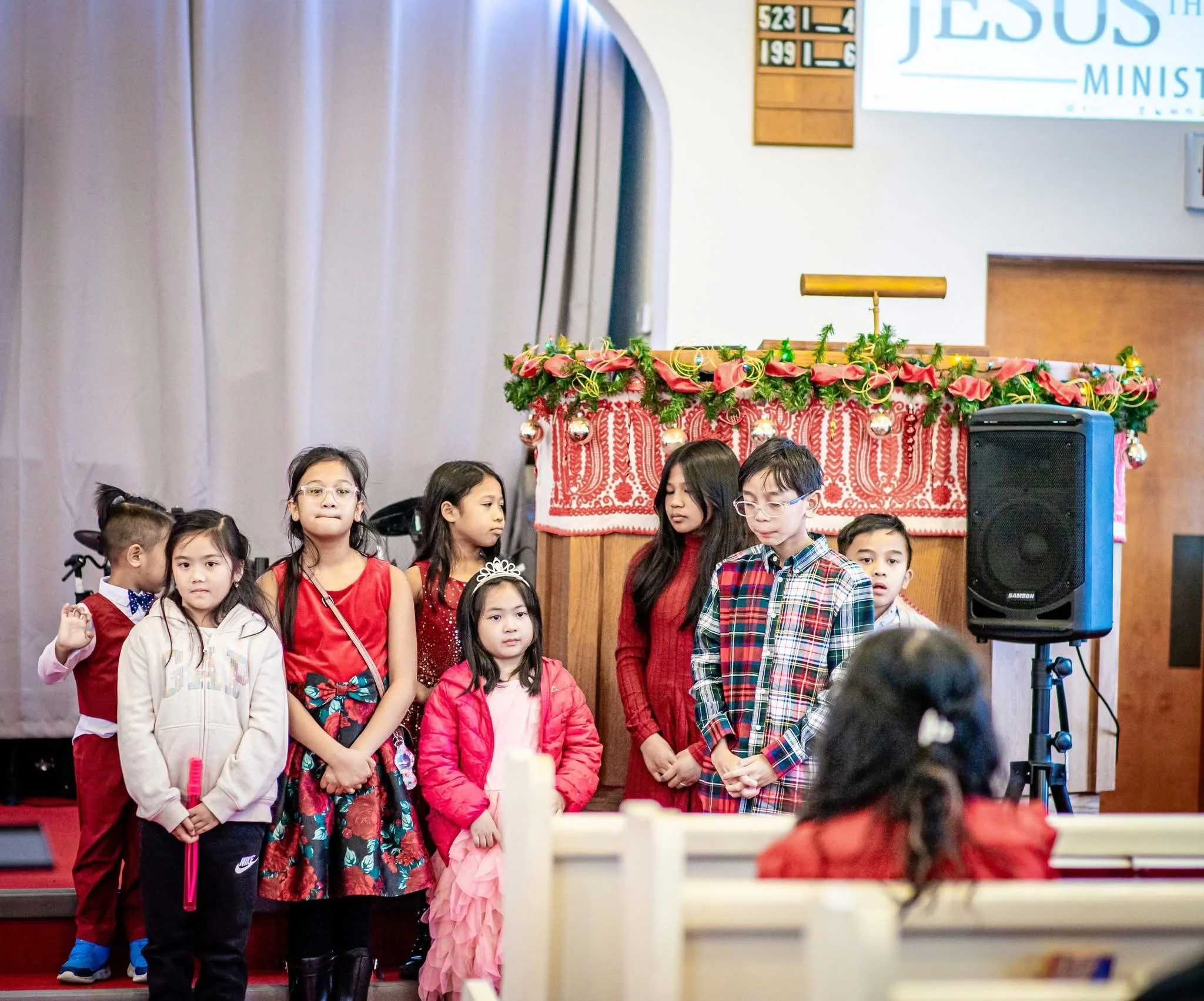 Children dressed in festive red and pink clothing stand on a stage decorated for Christmas, with a Christmas-themed backdrop and a Christmas tree.