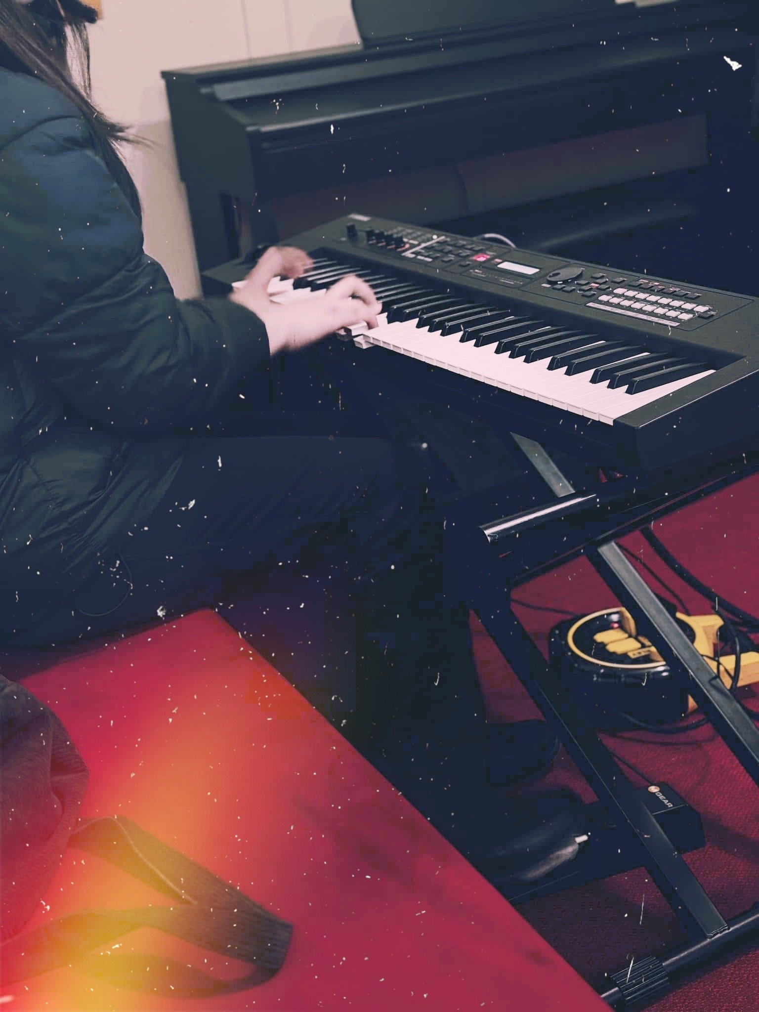 Person playing a black digital piano with a control panel, sitting on a red bench in a room with a red carpet.