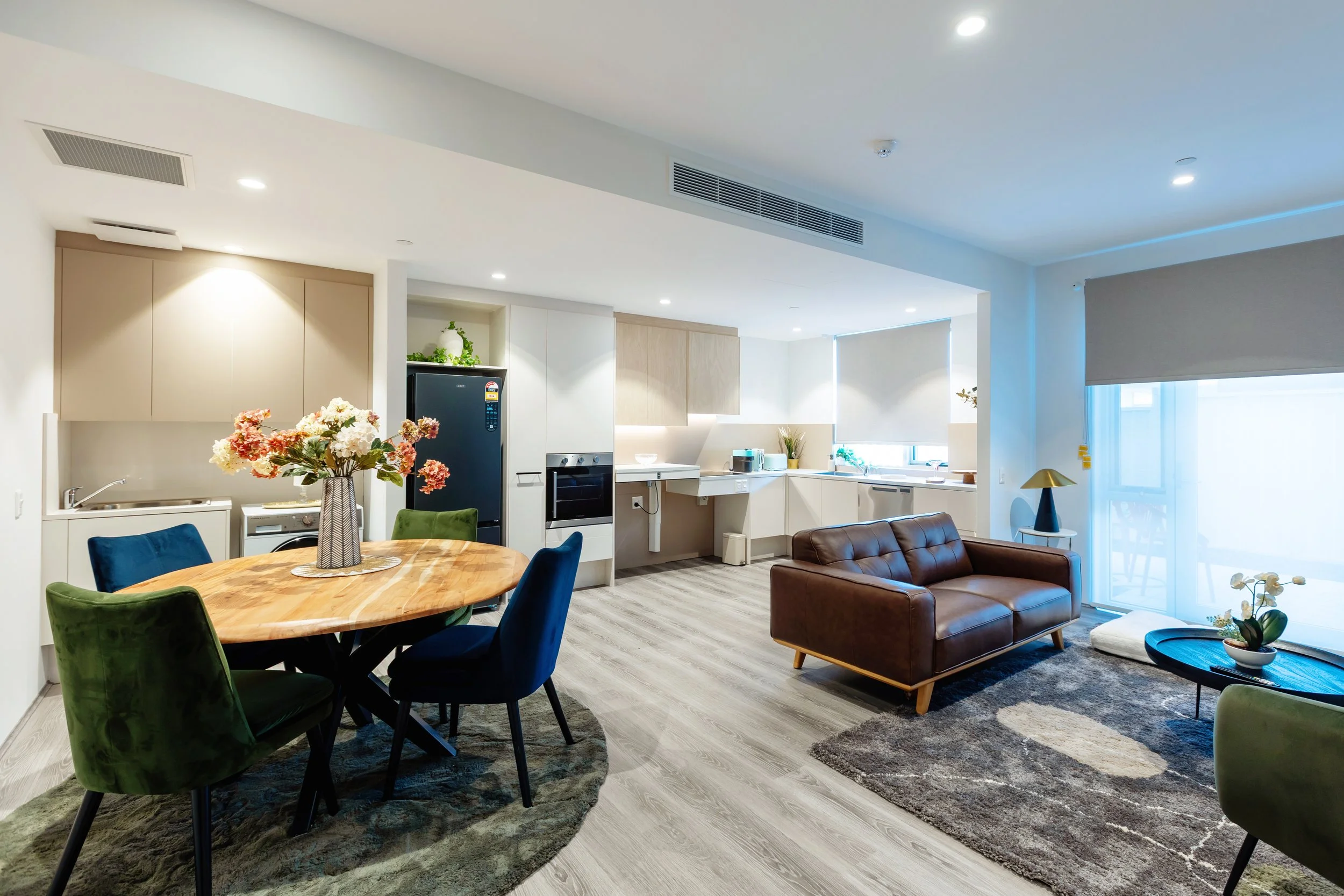 Modern open-concept living space with a kitchen, dining area, and sitting area with a brown leather sofa, a wooden dining table with colorful chairs, and a rug. Bright with natural light from large windows.