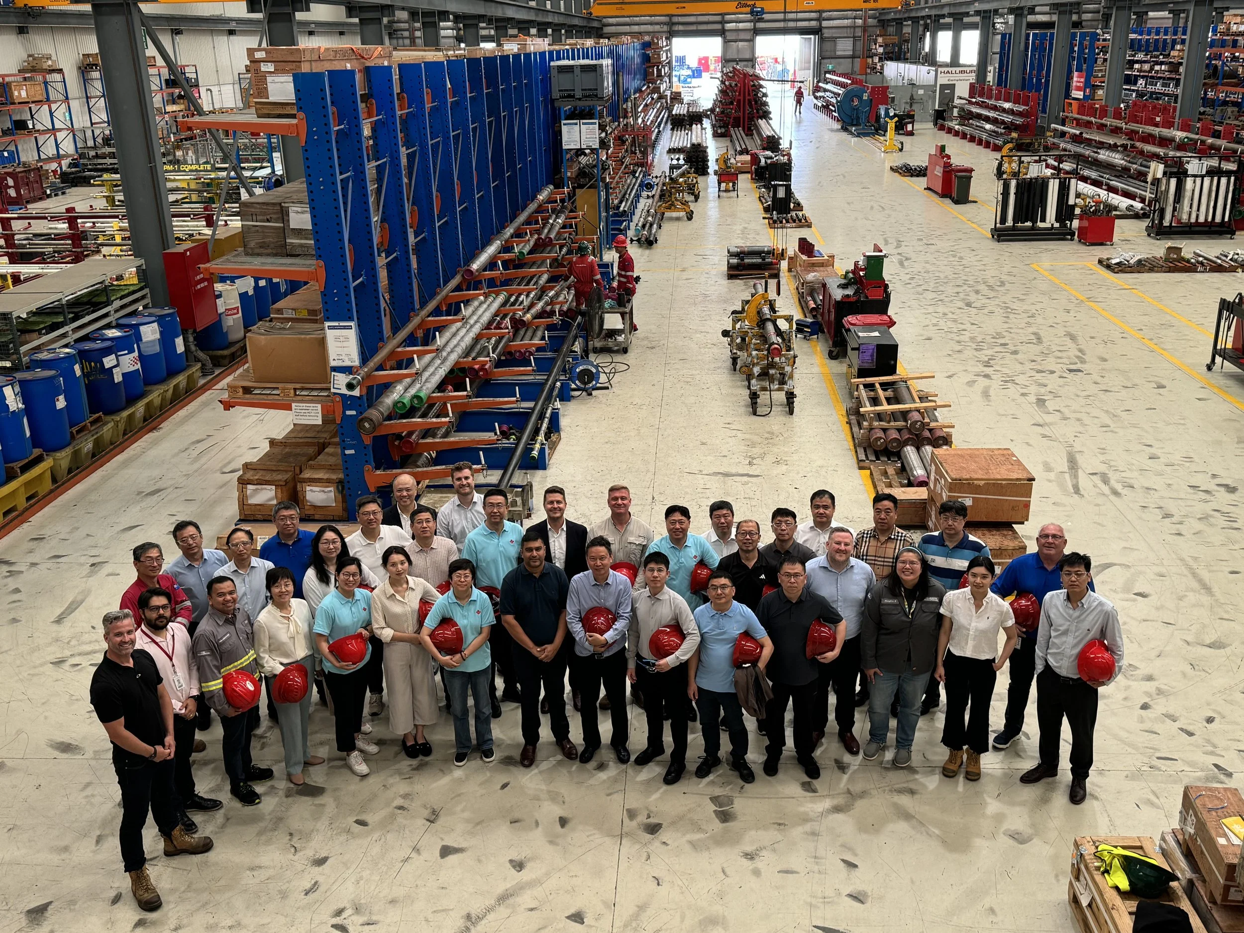 Group of factory workers and managers posing for a photo inside an industrial warehouse with shelves of materials, equipment, and machinery in the background.