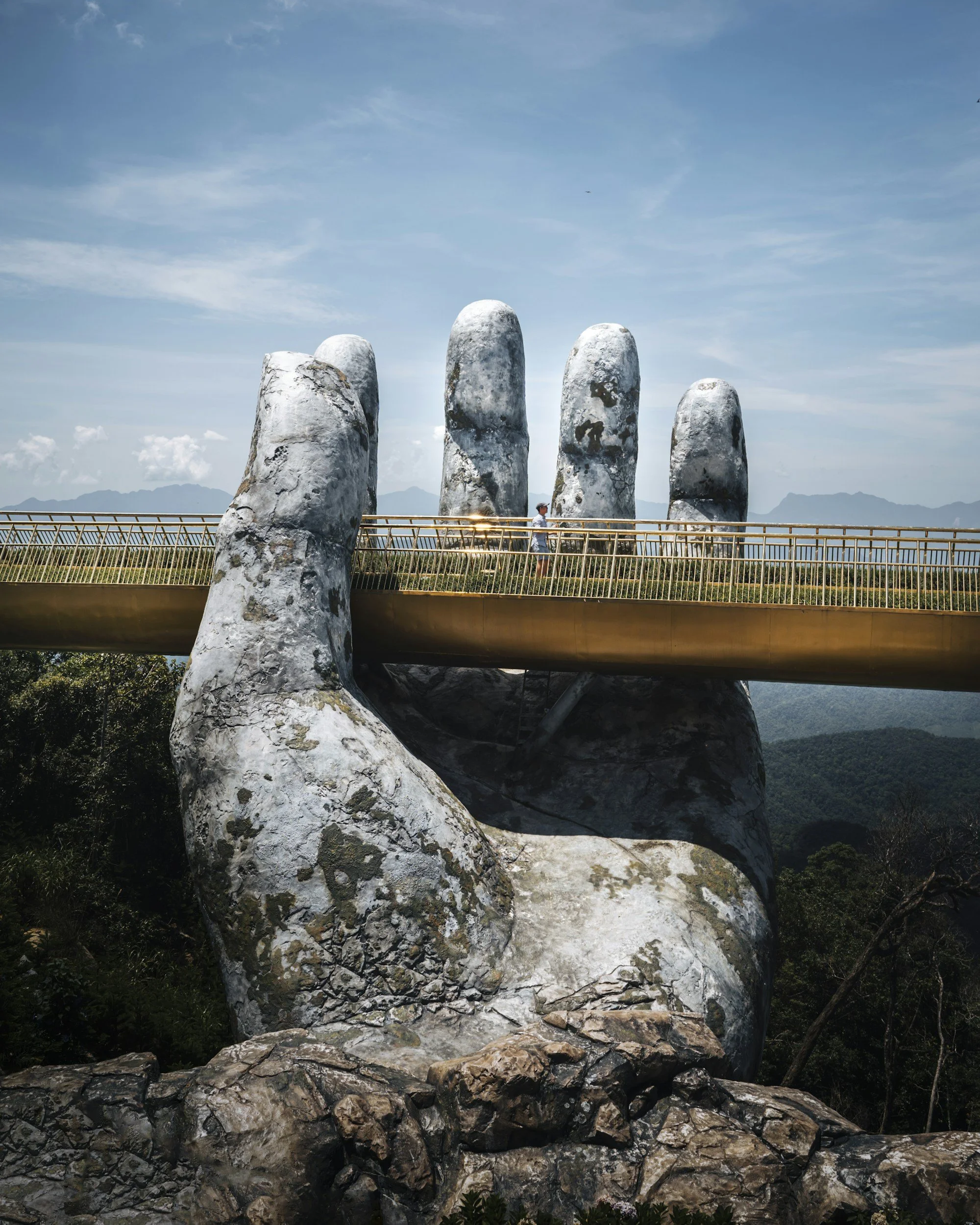 A large sculpture of a hand holding a bridge with five large stone fingers behind it, set against a blue sky with some clouds and mountains in the distance.
