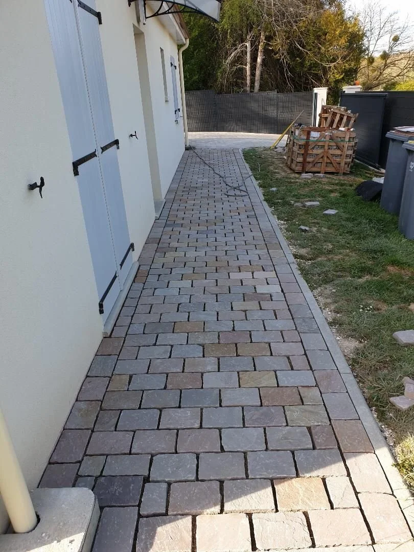 Une allée pavée devant une maison blanche, avec des bennes à ordures et un abri en bois, entourée de terrain en herbe et d'une clôture.