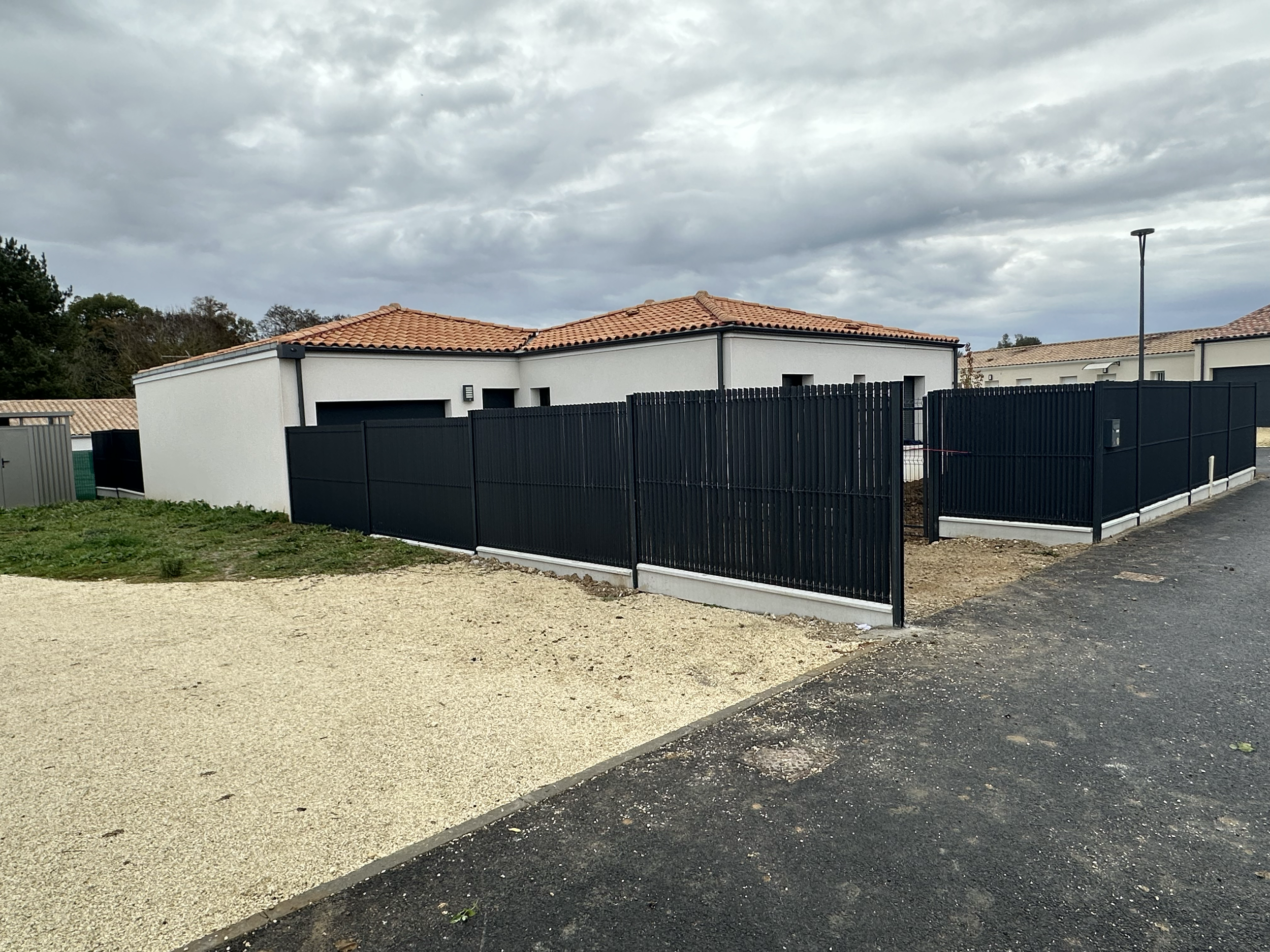 Une maison moderne avec toiture en tuiles rouges, entourée d'une clôture noire métallique, sous un ciel nuageux.