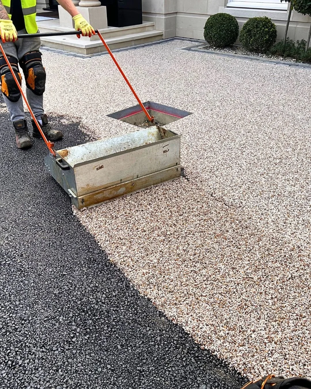 Travailleur en costume de sécurité appliquant un revêtement en résine sur un trottoir en pierre, créant une ligne entre deux sections de surface, devant une maison élégante avec des buissons taillés.
