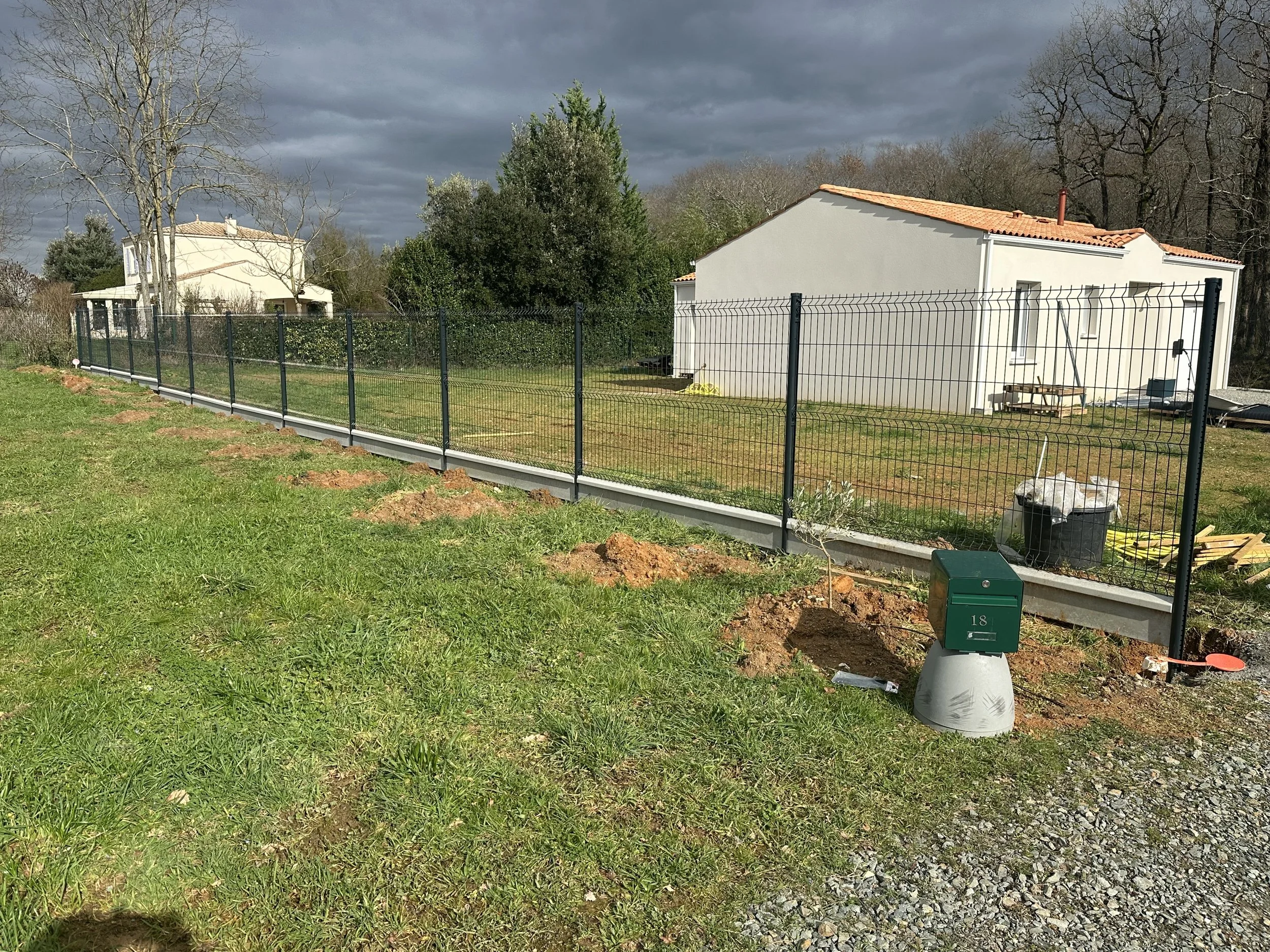 Clôture en métal noir entourant un petit jardin avec une maison blanche en arrière-plan, sous un ciel nuageux.
