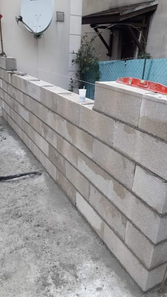Mur en blocs de béton en construction avec une tasse en haut, un niveau à bulle rouge, et une plaque satellite sur le mur à gauche.