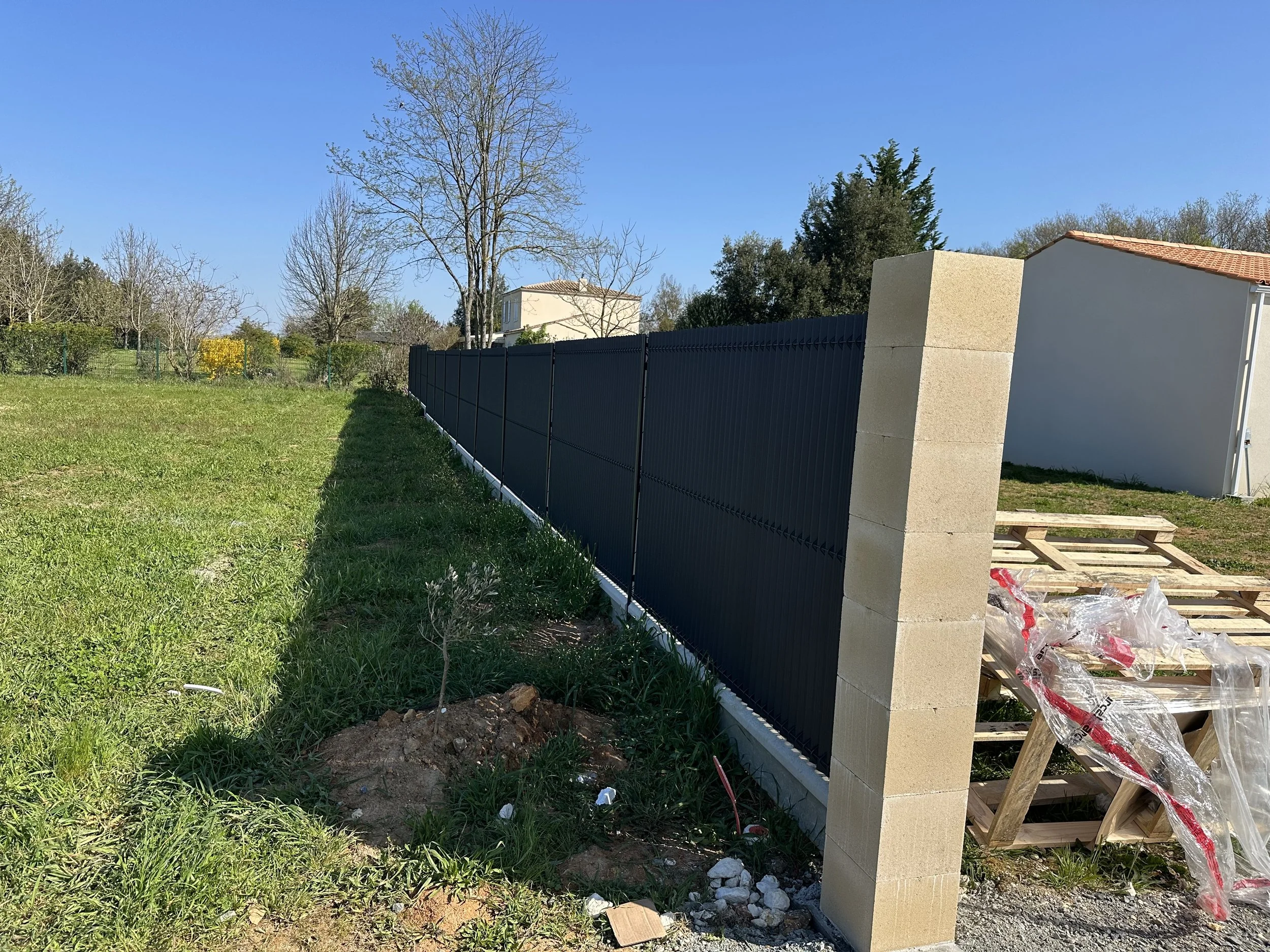 Mur de clôture noir en métal avec nouveau pilier en pierre beige dans un jardin vert sous ciel bleu