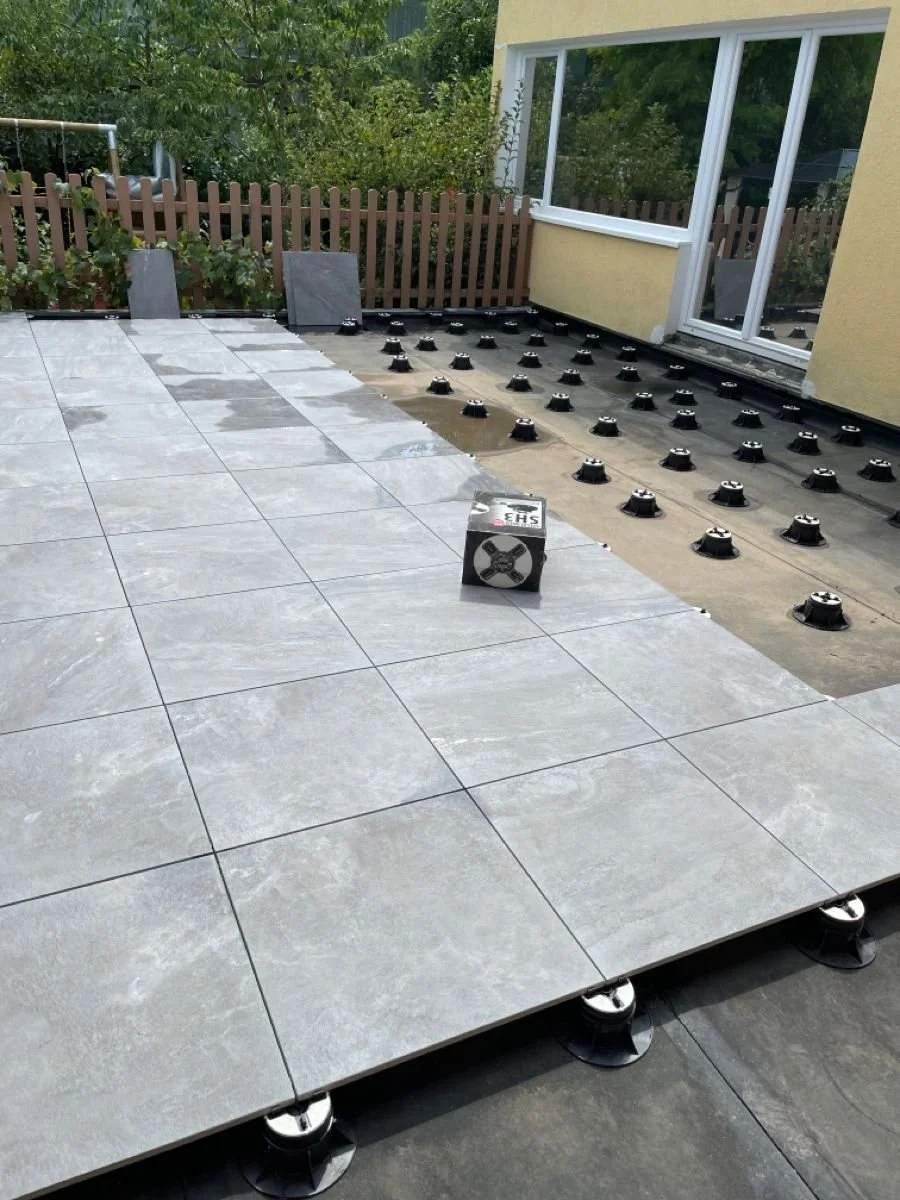 Installation de tuiles sur une terrasse avec des plots de nivellement et un carton de matériel. Fenêtre et jardin en arrière-plan.