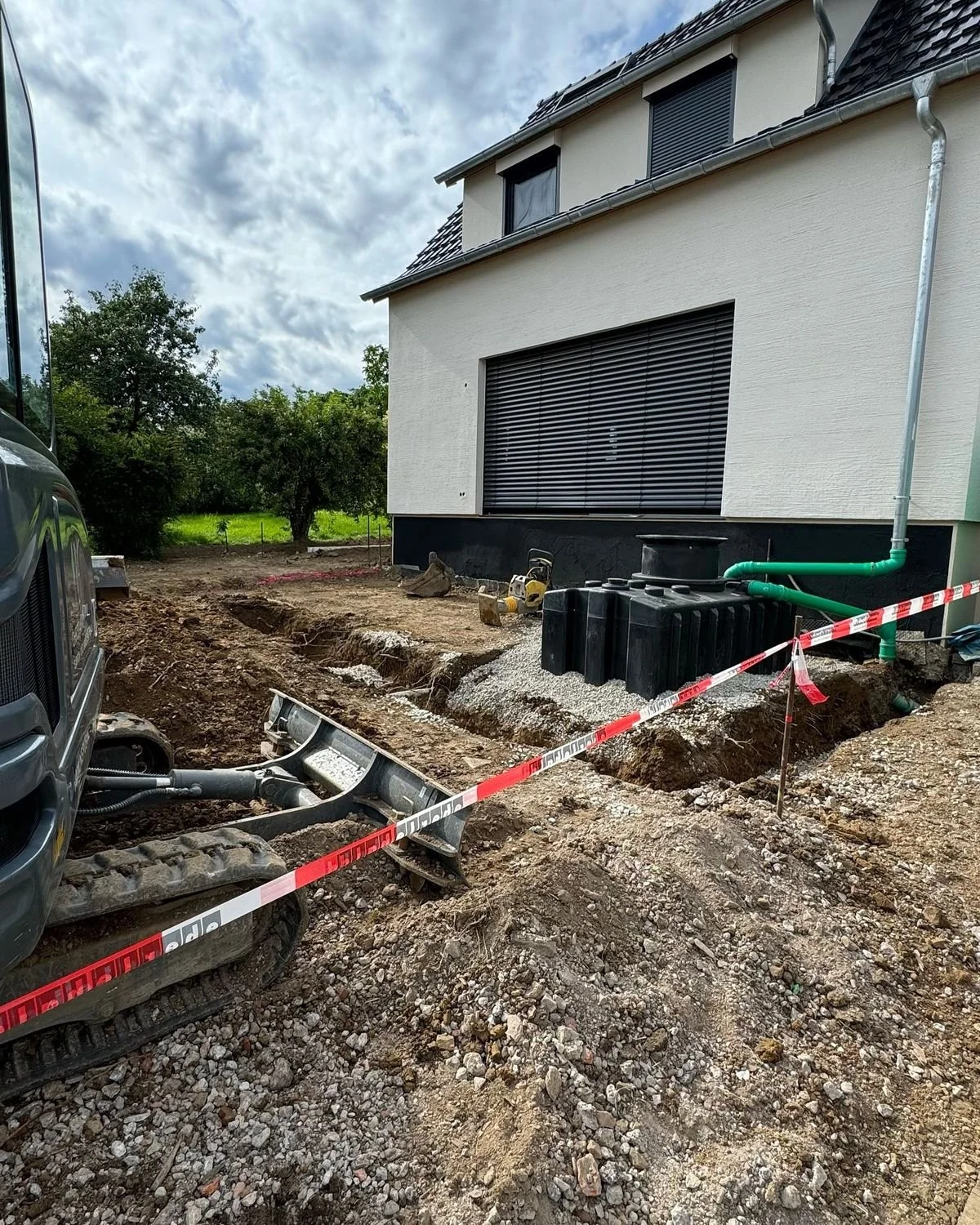 Travaux de construction autour d'une maison, avec une excavation dans le sol, une pelle mécanisée, et un système d'évacuation d'eau, en attente d'installations ou de rénovations.
