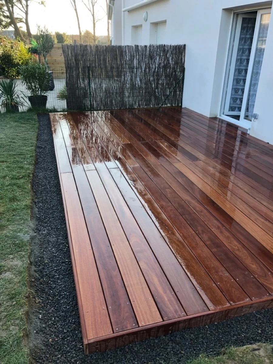 Terrasse en bois tout en rainures, récemment lavée, située à l'arrière d'une maison avec une baie vitrée et un mur blanc.