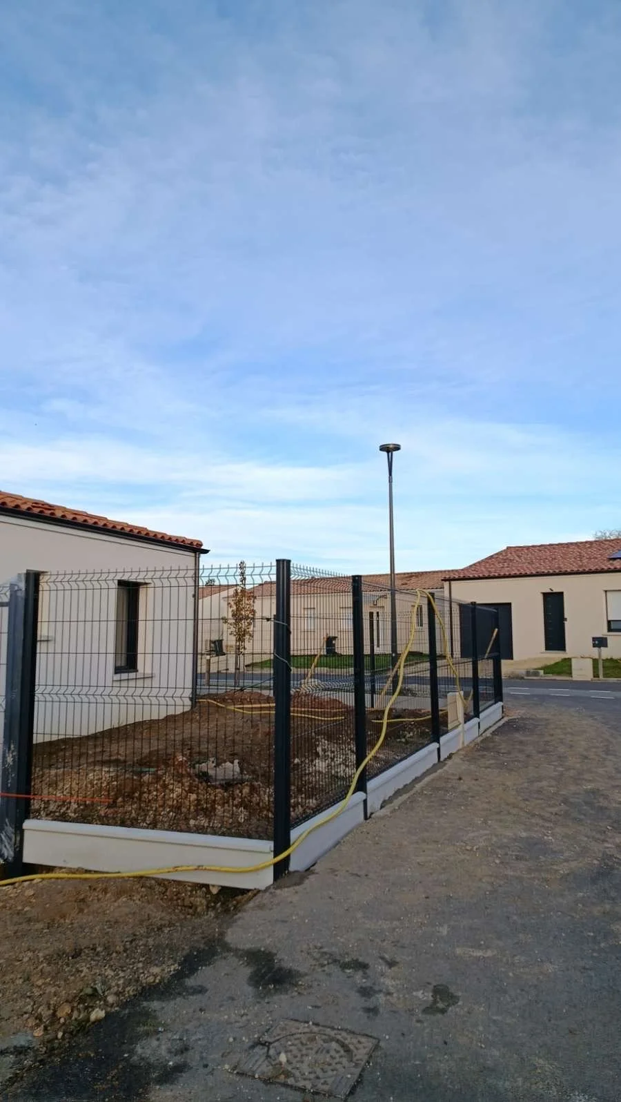 Un espace en cours d'aménagement avec une clôture noire, un arbre, un lampadaire, et des bâtiments résidentiels en arrière-plan sous un ciel bleu.