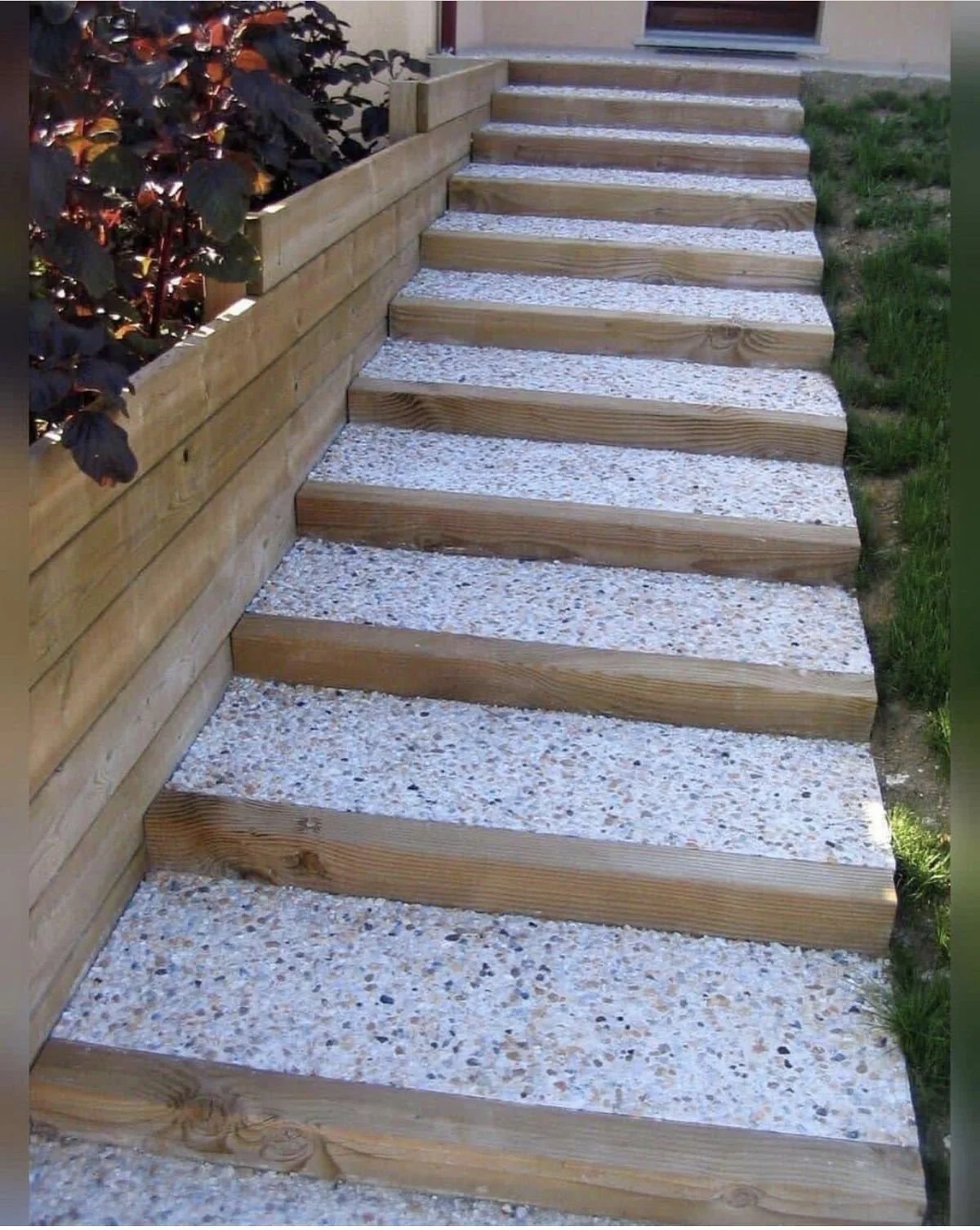 Escalier en bois avec du gravier entre chaque marche, bordé par une clôture en bois et un massif de plantes à gauche.