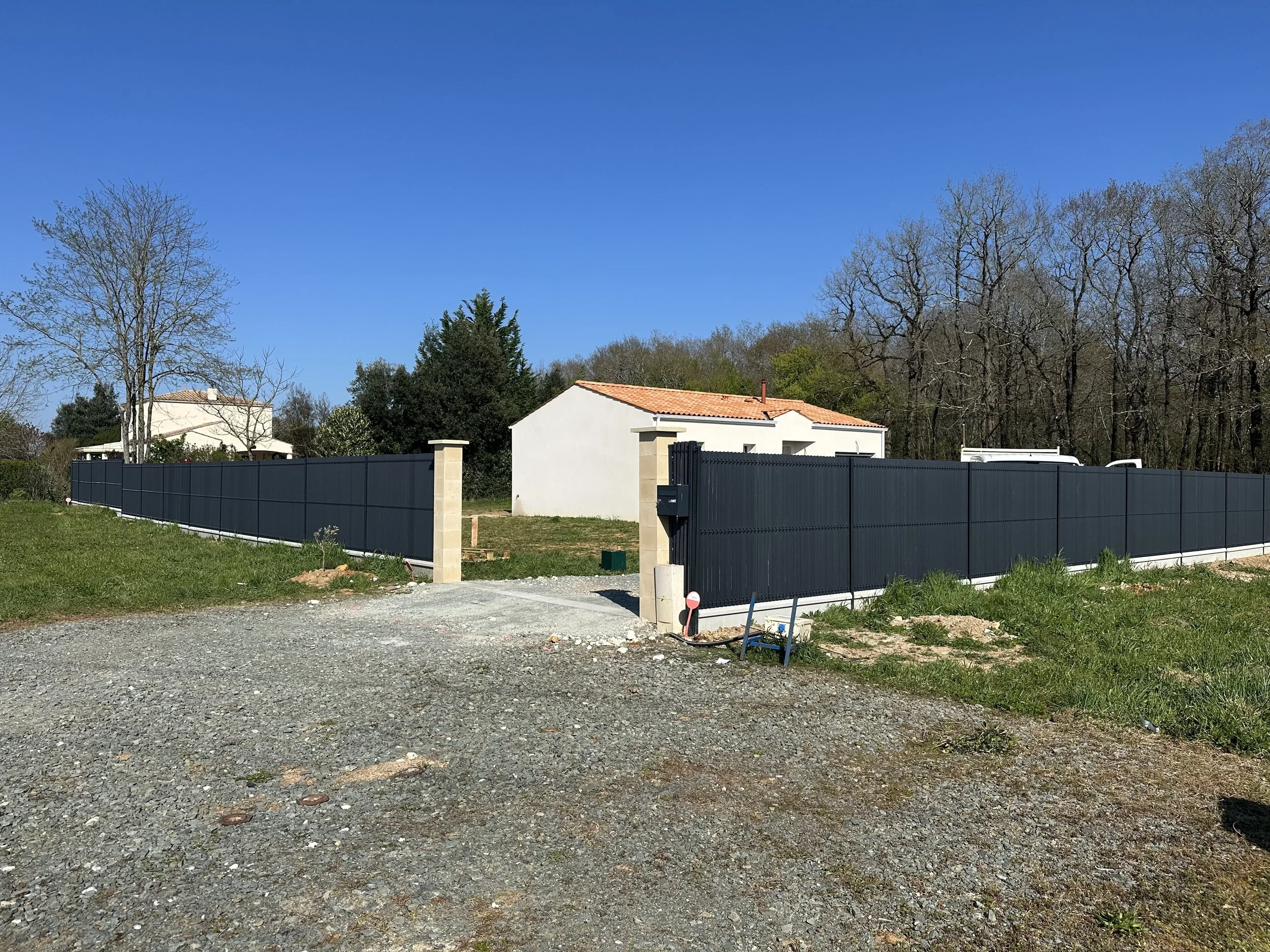 Une clôture noire entourant une maison blanche avec un toit en tuiles, à l'extérieur d'une zone de construction ou d'aménagement, avec un ciel bleu clair et des arbres en arrière-plan.