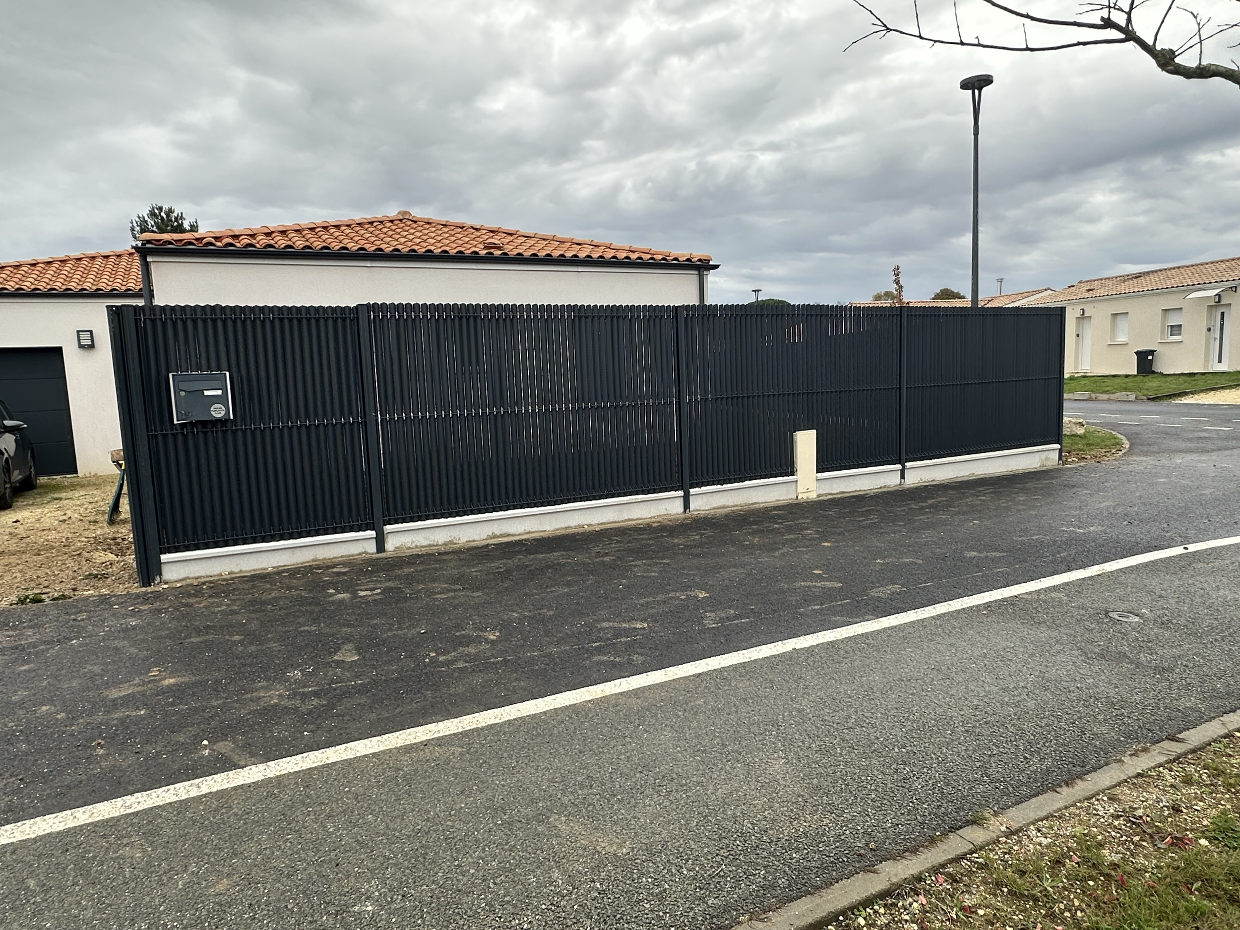 Clôture en métal noire entourant une maison résidentielle, avec boîte aux lettres, sous un ciel nuageux.