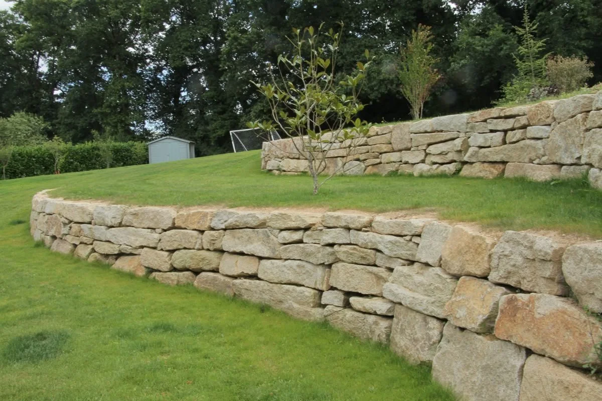 Mur en pierre avec plusieurs niveaux de terrasses recouvertes d'herbe verte, dans un jardin avec des arbres en arrière-plan.