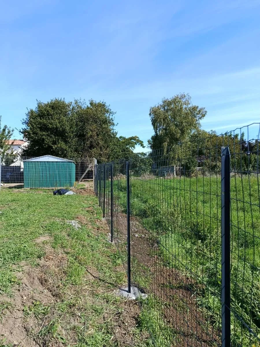 Une clôture métallique noire délimite un terrain avec de l'herbe verte. Au fond, il y a un petit cabanon vert, quelques arbres et un ciel bleu.