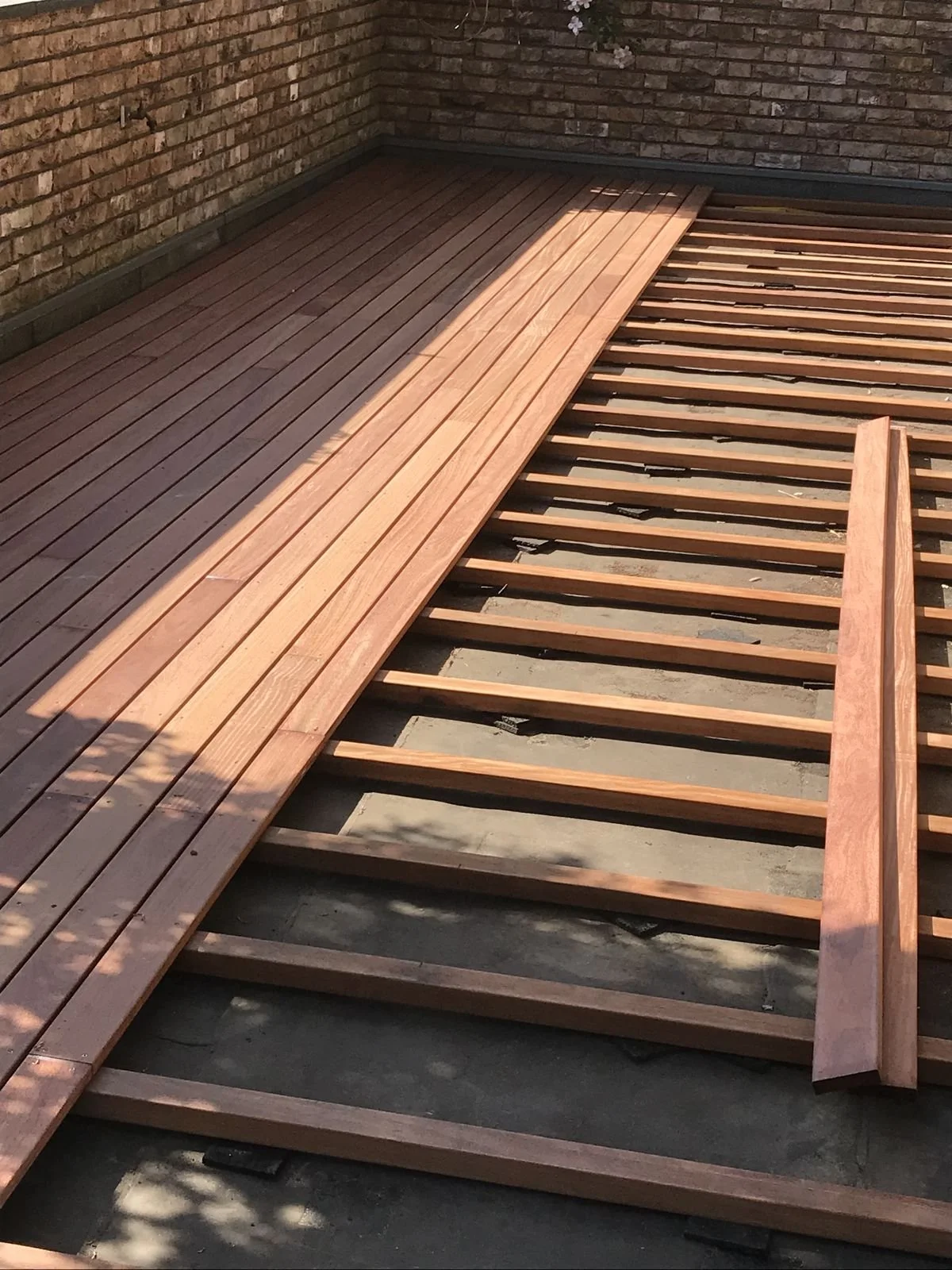 Une terrasse en cours de construction avec des planches de bois posées sur une structure de support. La moitié gauche de la terrasse est déjà couverte de planches en bois. La terrasse est adjacente à un mur en briques.