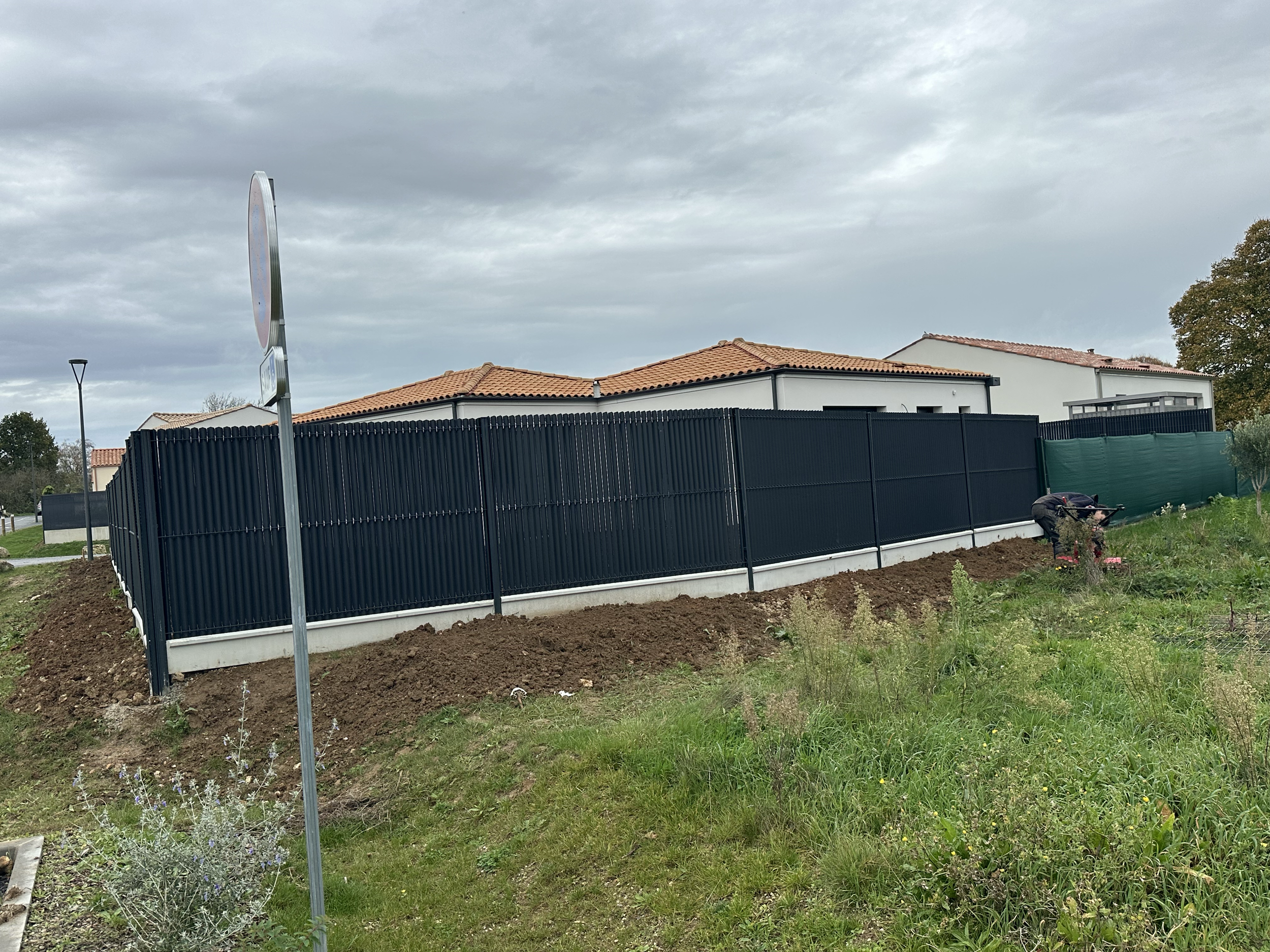 Muret en panneaux de clôture noirs, maison avec toiture en tuiles rouges, et une personne travaillant dans un jardin à côté.