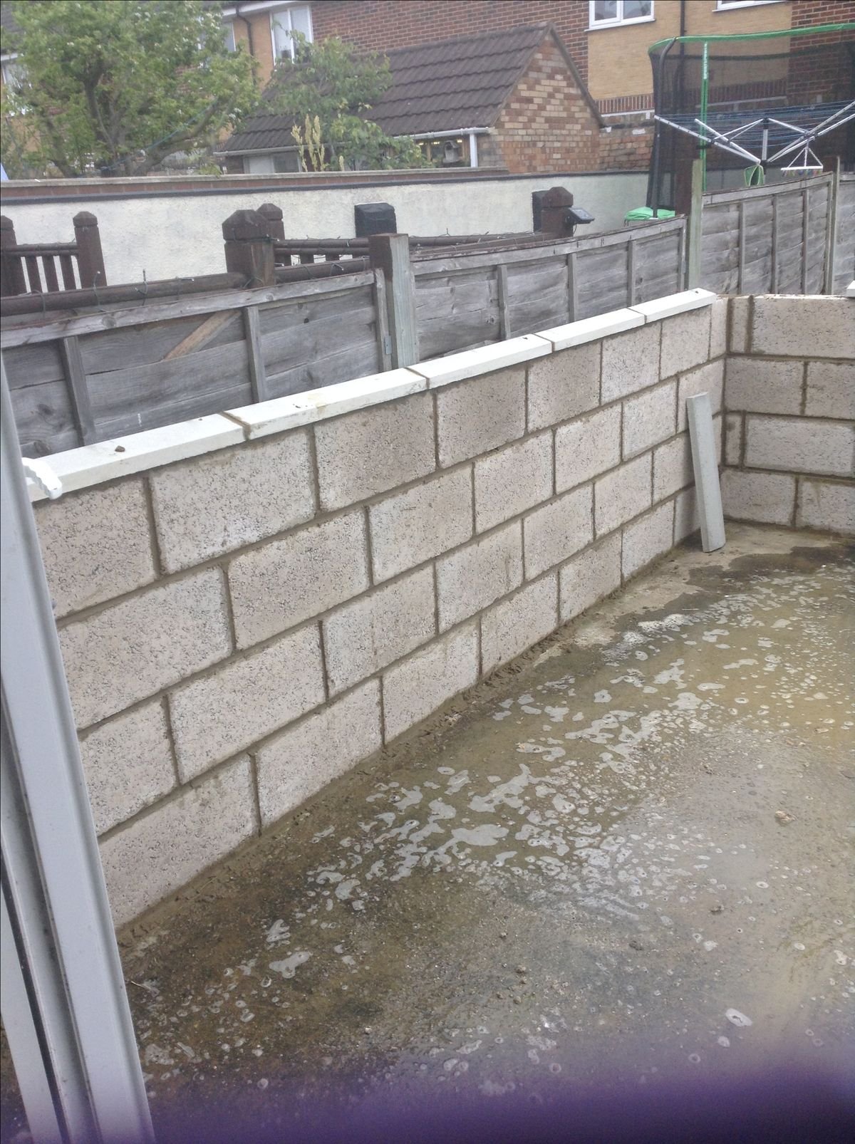 Un balcon en construction avec un mur en blocs de béton, sol bétonné avec des traces d'eau, et un bâtiment résidentiel en arrière-plan.