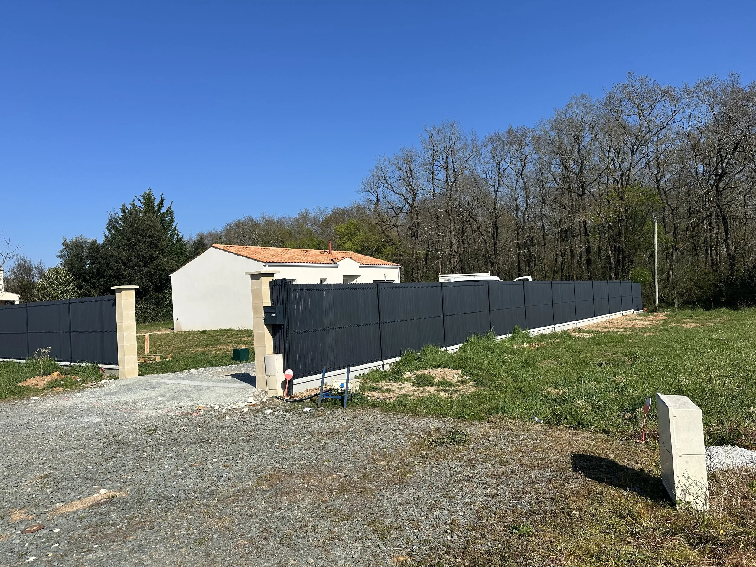 Vue d'une clôture en métal gris foncé autour d'une maison blanche avec un toit en tuiles, sous un ciel bleu clair, avec des arbres sans feuilles en arrière-plan et un sol partiellement en gravier et en herbe.