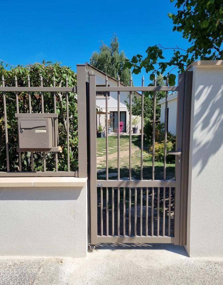 Une porte en métal grise encadrant une clôture, avec un jardin derrière, comprenant des arbres, des buissons et une maison en arrière-plan, sous un ciel bleu clair.