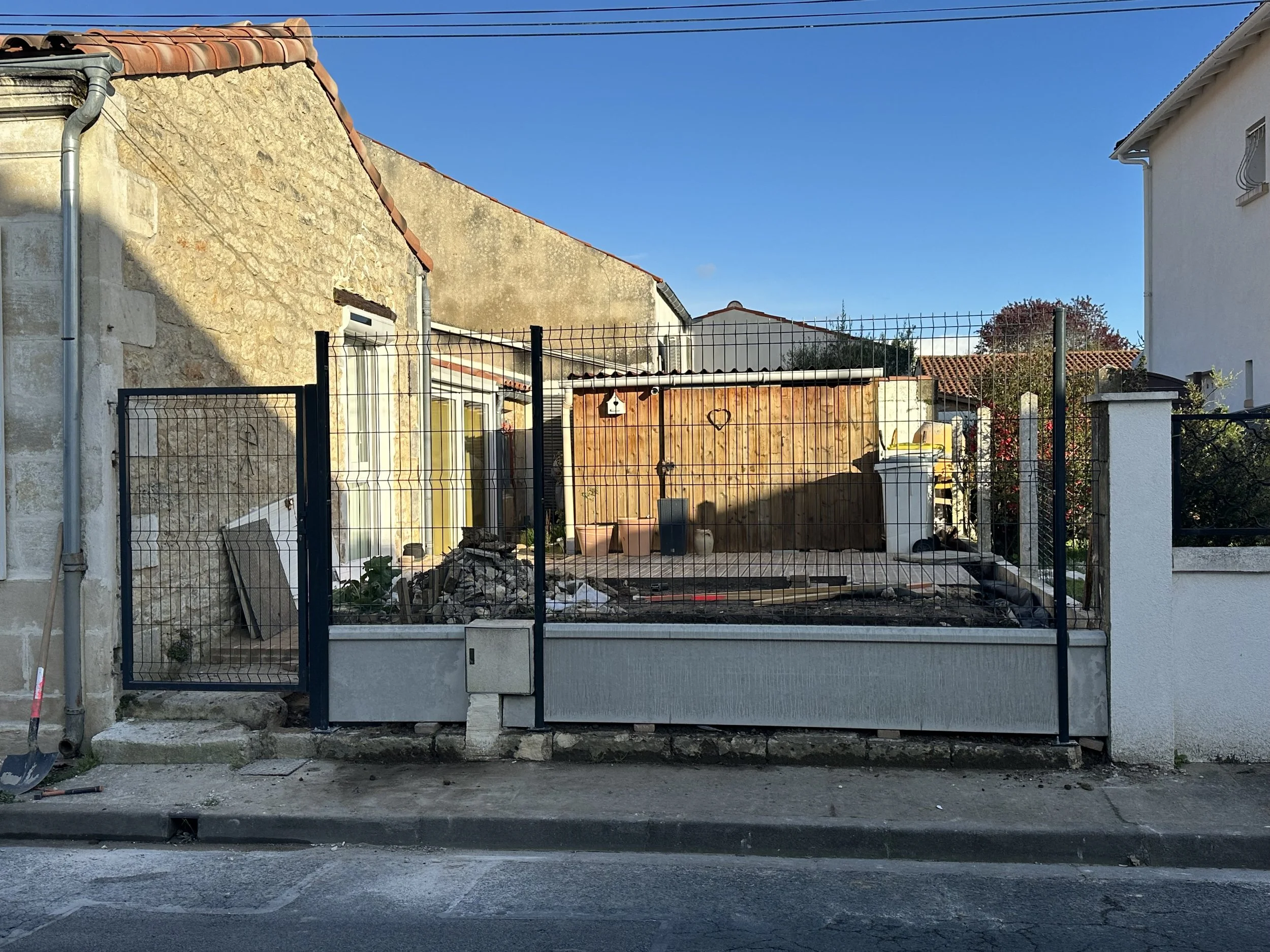 Cour clôturée avec chantier de construction, matériel et piles de déblais, à côté d'une maison en pierre, sous un ciel bleu.