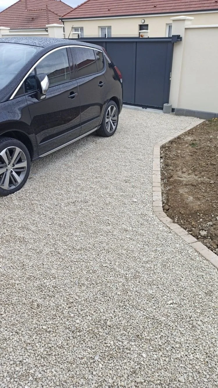 Une voiture noire garée sur une allée en gravier en face d'une porte de clôture en métal, avec une maison en arrière-plan.