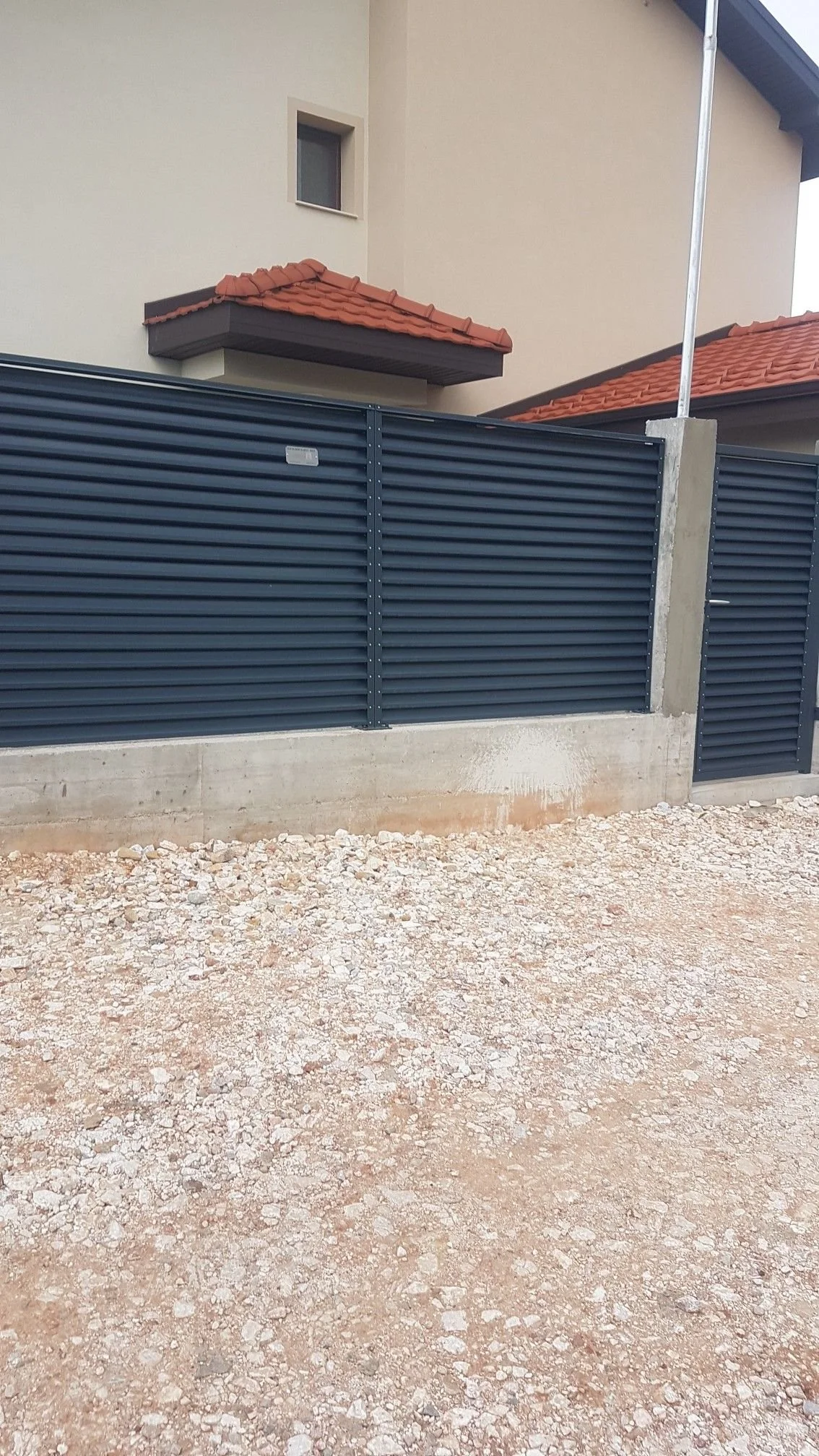 Une clôture en métal bleue devant une maison avec un mur en béton, un portail et un toit en tuiles rouges. Le sol est en terre gravelée.