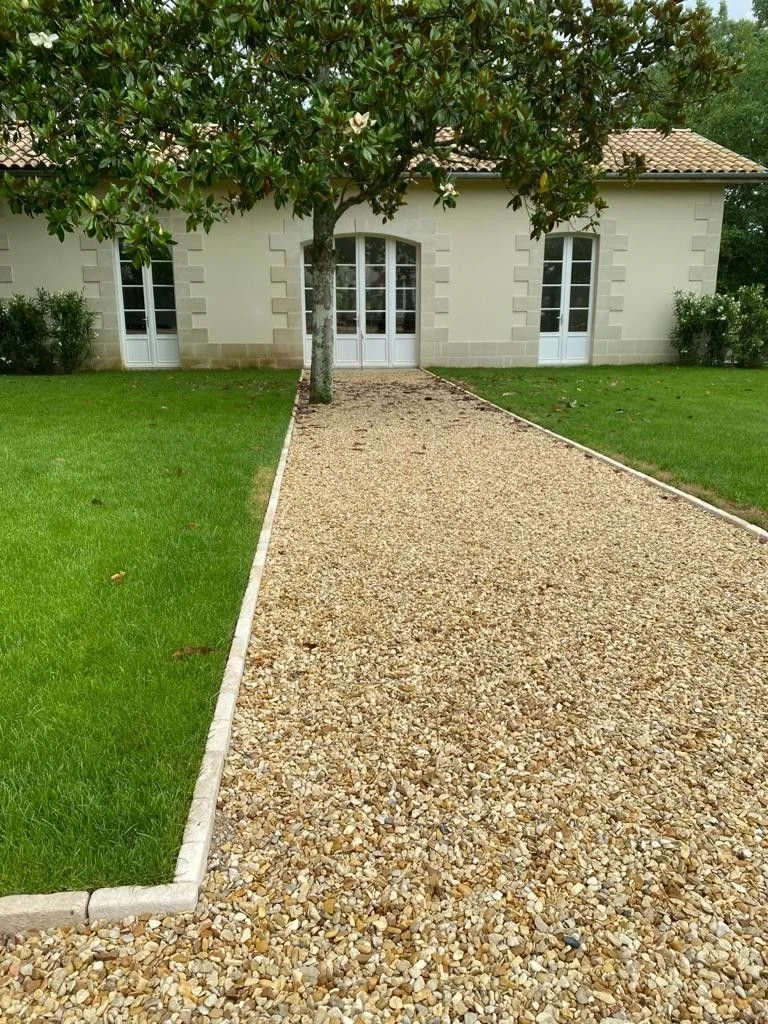 Chemin en gravier menant à une maison avec des murs en pierre et des fenêtres en verre, ombragé par un arbre au centre du chemin, avec des zones de pelouse verte des deux côtés.