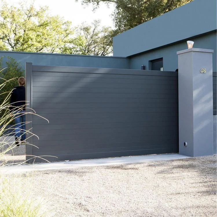 Une clôture en métal gris foncé devant une maison moderne avec une porte automatique. Il y a une personne à gauche, en train d'utiliser la porte. À droite, un pilier avec un numéro 666.