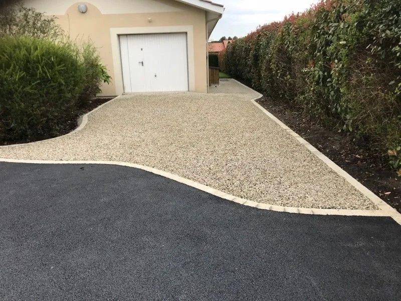Allée de voiture en enrobé noir et gravier menant à un garage avec un mur végétal sur le côté droit et un jardin avec des arbustes à gauche.