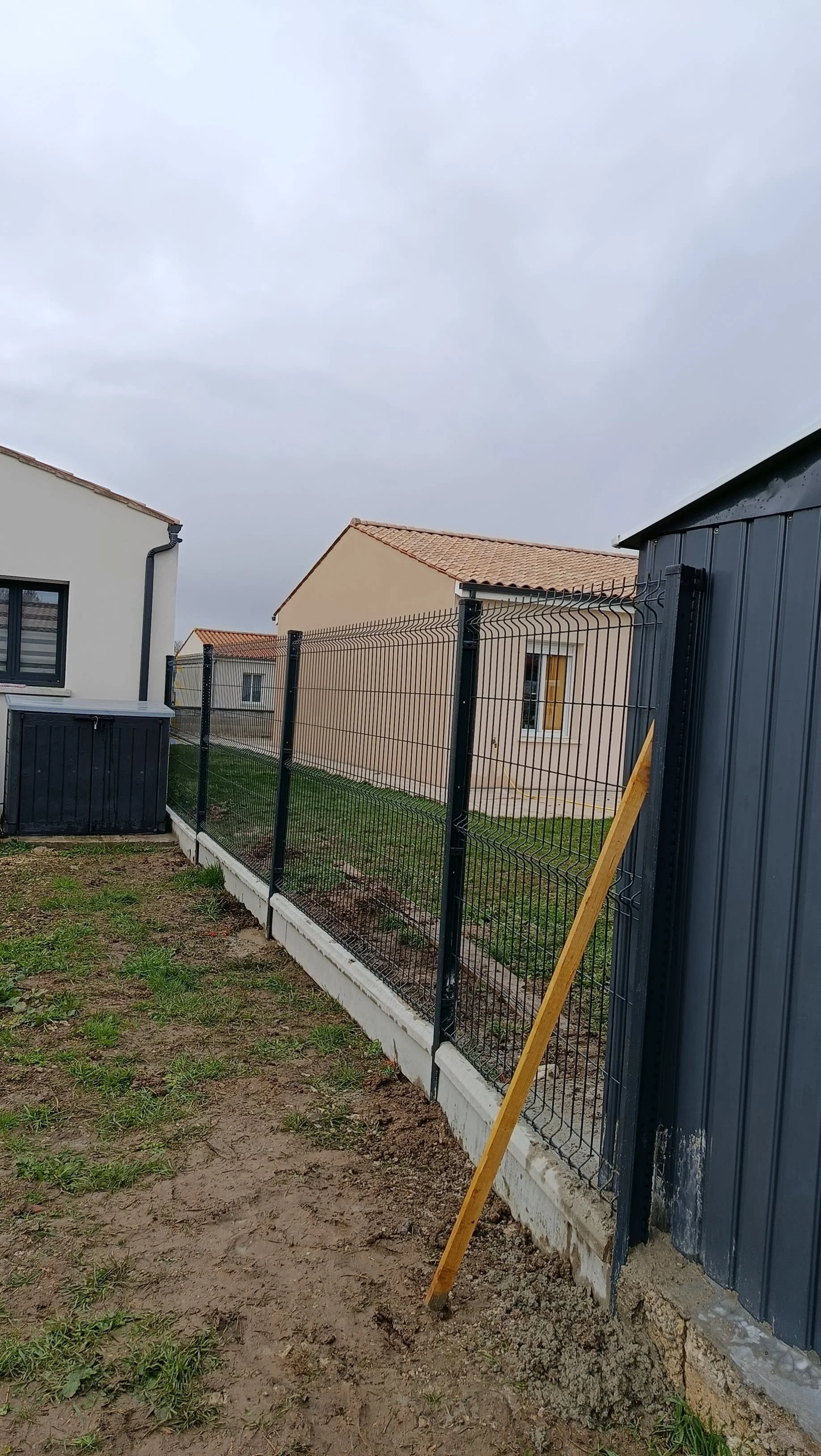 Clôture en cours d'installation dans un jardin de maison, avec de petites maisons en arrière-plan, ciel nuageux.