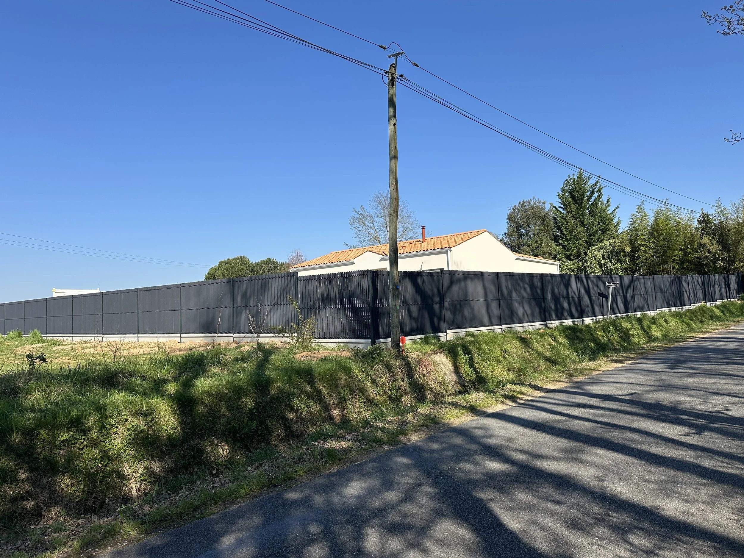 Une maison avec un toit en tuiles oranges derrière une clôture noire, un poteau électrique en bois, un chemin en gravier, du soleil et un ciel bleu clair.