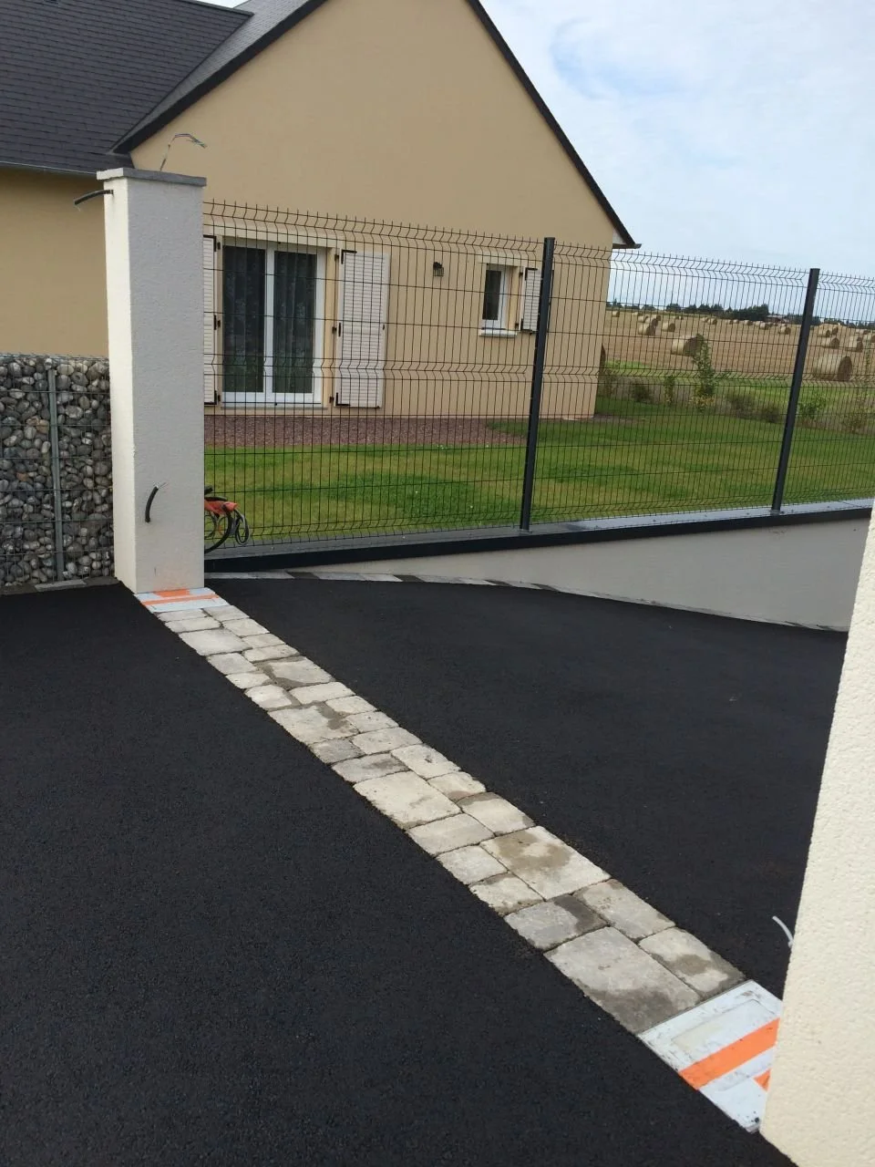 Voie d'accès pour voiture avec pavés, menant à une maison avec un portail en métal et un jardin.