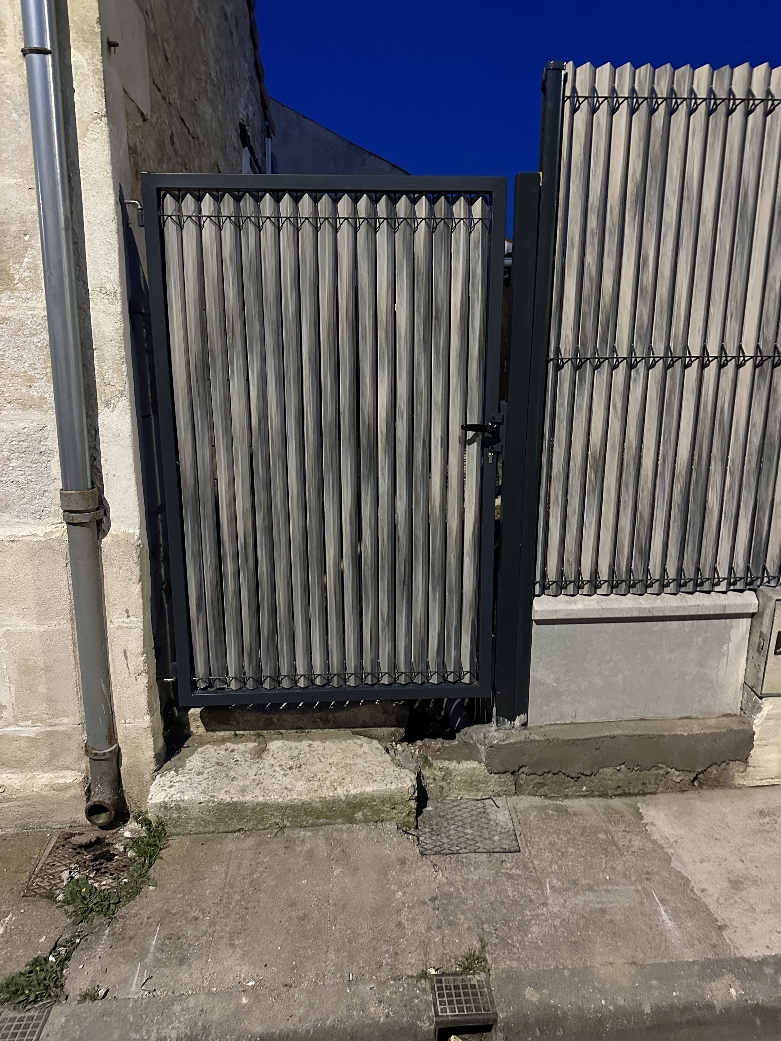 Une porte en métal gris avec des barres verticales en bois, fixée à un mur en pierre, sous un ciel nocturne. La porte est équipée d'une serrure et se trouve sur un trottoir en béton avec quelques plantes poussant dans la fissure
