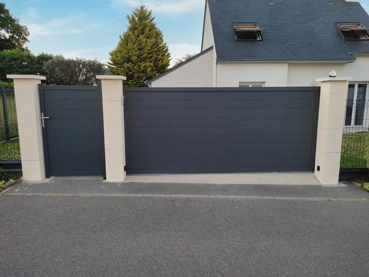 Panneau de clôture en métal gris foncé avec deux piliers en pierre blanche, séparant une maison résidentielle et une allée