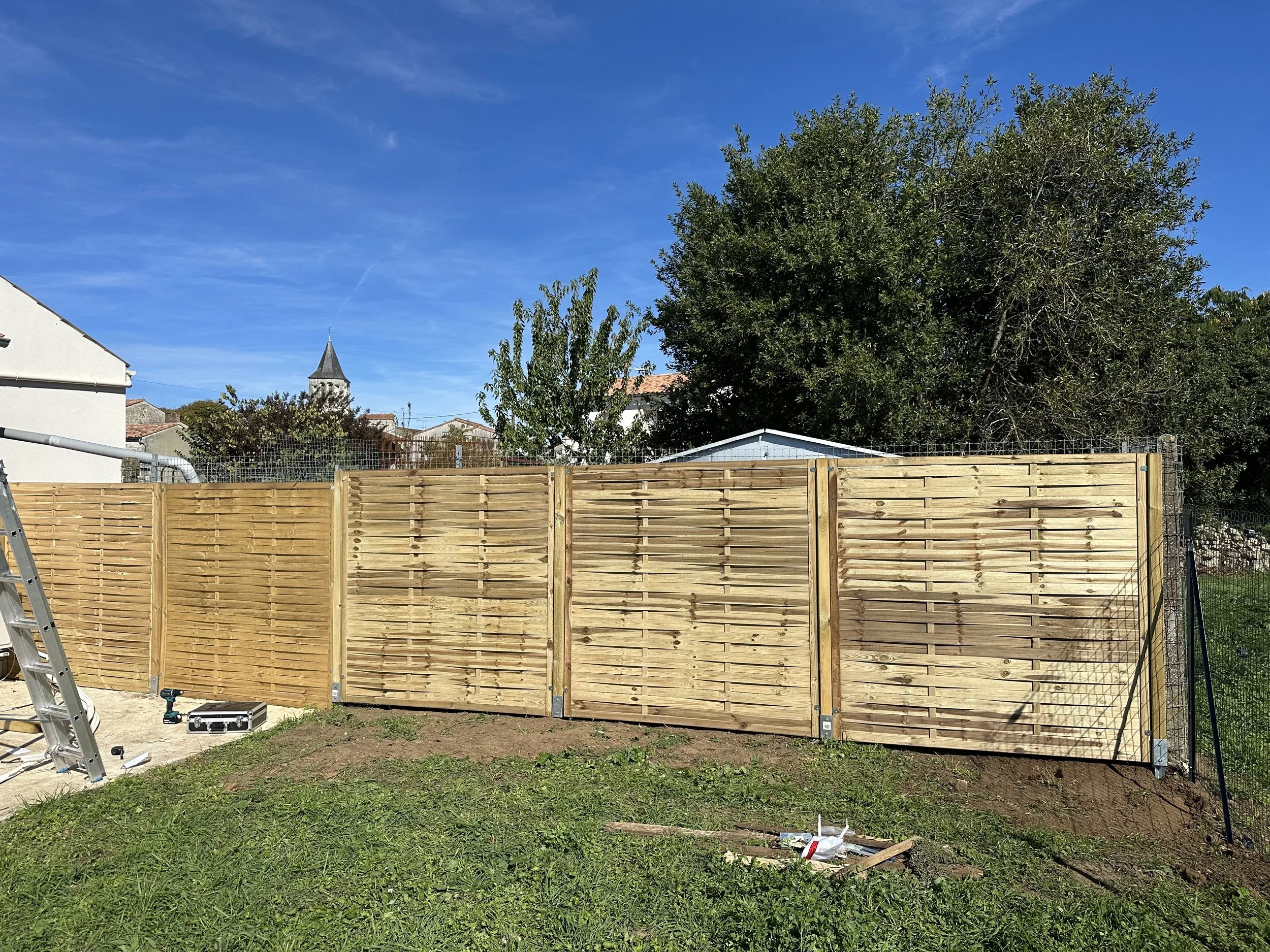 Nouveau panneau de clôture en bois en construction dans un jardin avec un ciel bleu et un arbre en arrière-plan.