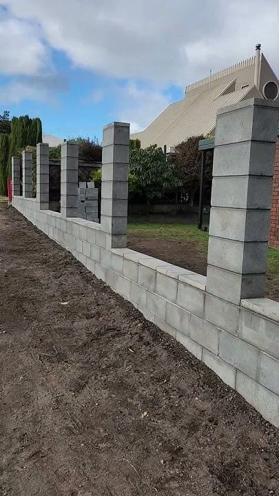 Mur en construction avec des blocs de béton sur un terrain de terre, avec un bâtiment moderne en arrière-plan.