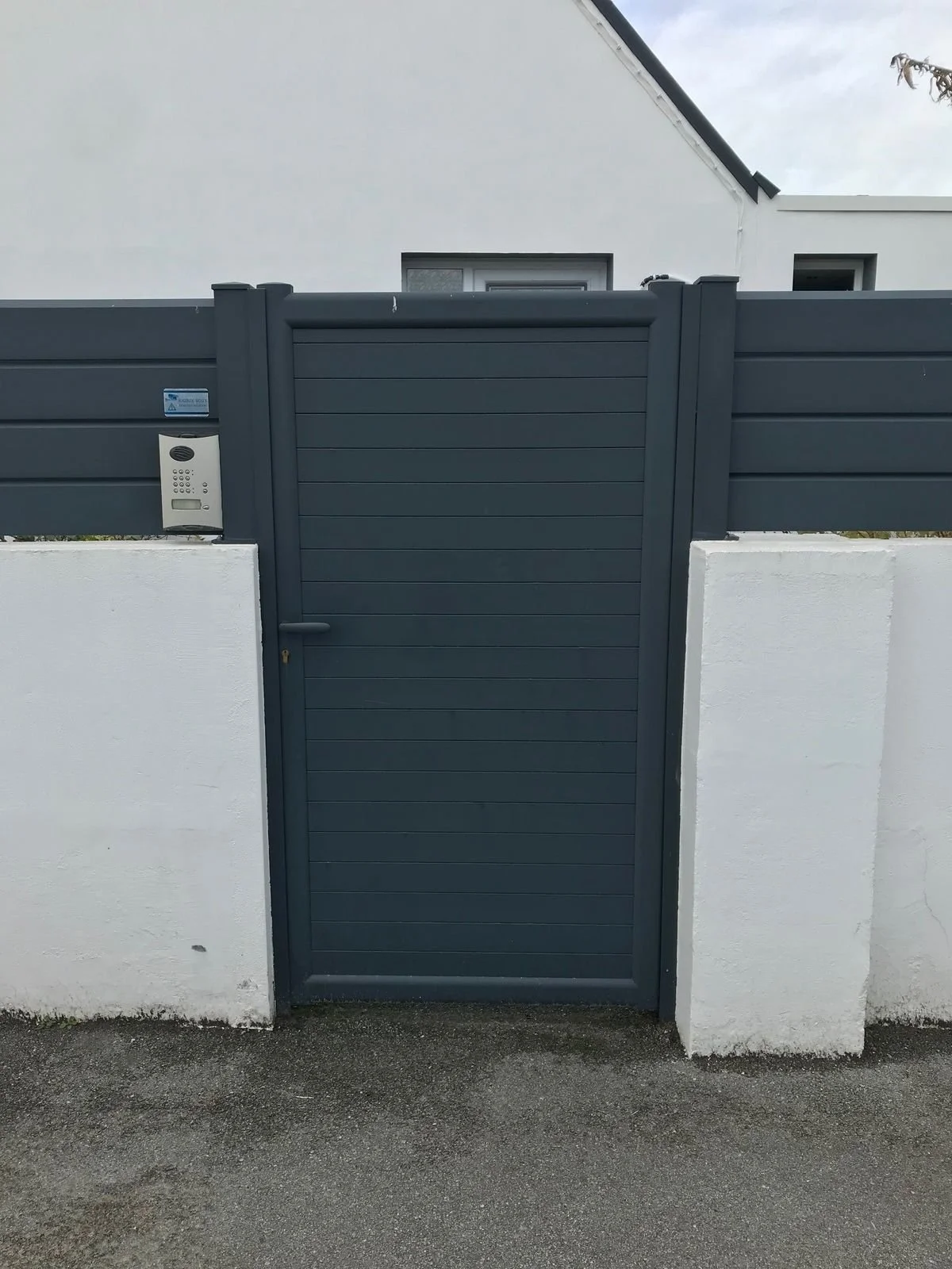 Une porte en bois ou en matériau composite de couleur grise, encadrée par un mur blanc et un panneau de clôture gris, avec un interphone fixé sur le mur à gauche.